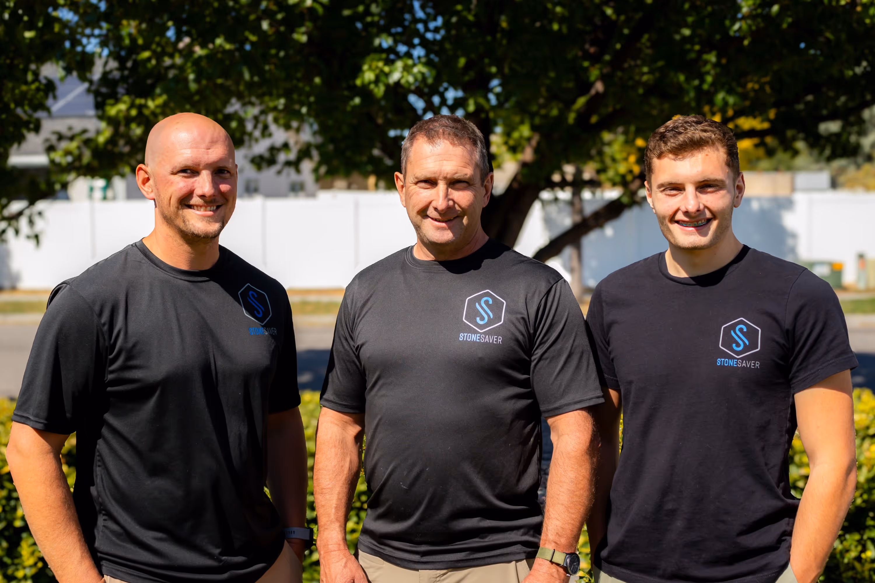 Three men wearing black StoneSaver shirts posing outdoors with trees and white fence in background.