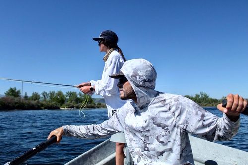 Man wearing a hood and sunglasses while rowing a drift boat down the Bighorn River in Montana. A girl stands in the back holding a fly fishing rod.