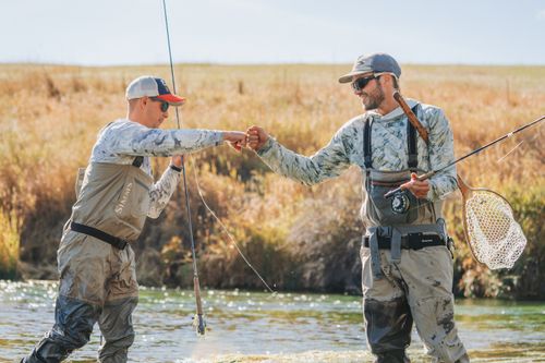 Two guys in waders standing in a river fist bump after catching a fish.