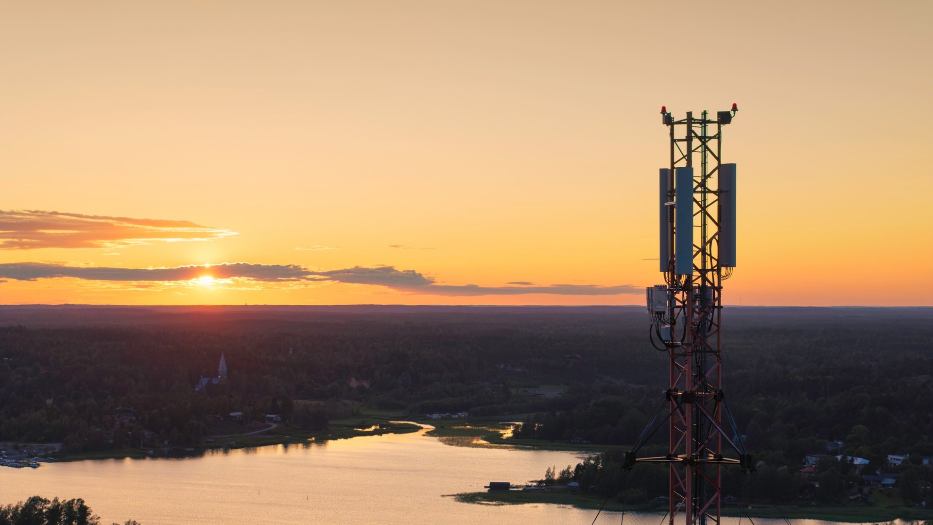 Cell tower with 5G antennas above a lake and forest at sunset
