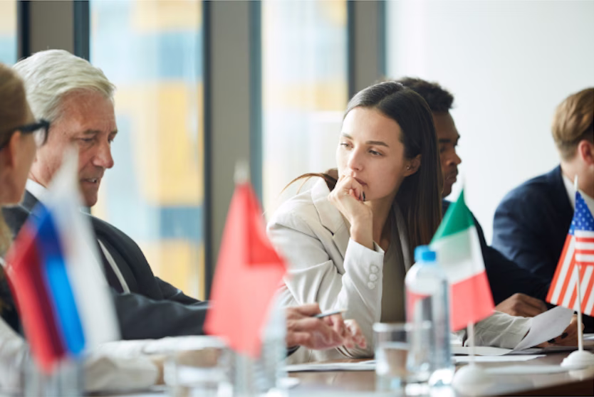 A diverse group of governance professionals at an international meeting table with country flags, with a woman in a contemplative pose representing strategic deliberation under uncertainty