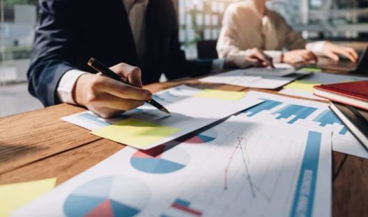 Two professionals reviewing data charts and reports spread across a table, representing the challenge of making sense of complex external signals in enterprise strategy and intelligence workflows.