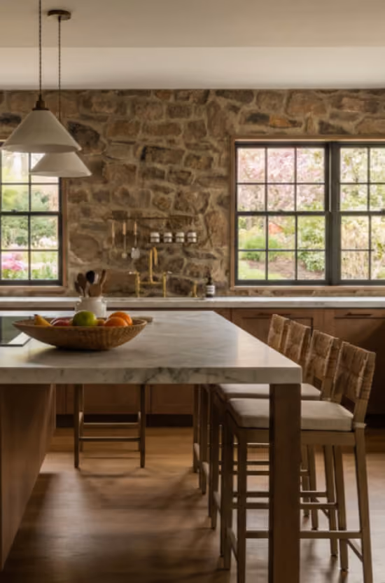 Rustic kitchen with stone wall, marble island, hanging lamps, wooden bar stools, and large windows.