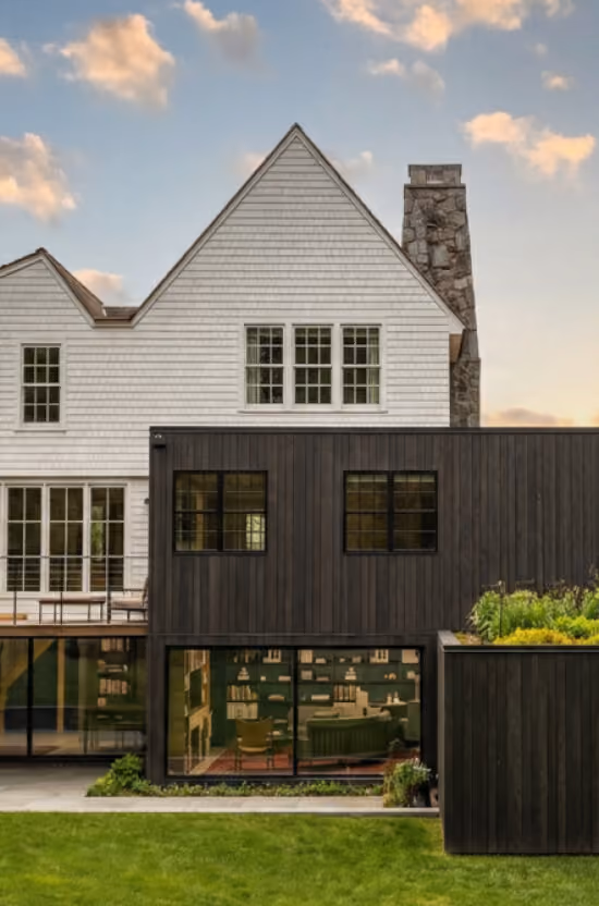 Modern two-story house with white upper siding, dark lower wood paneling, stone chimney, and green lawn.