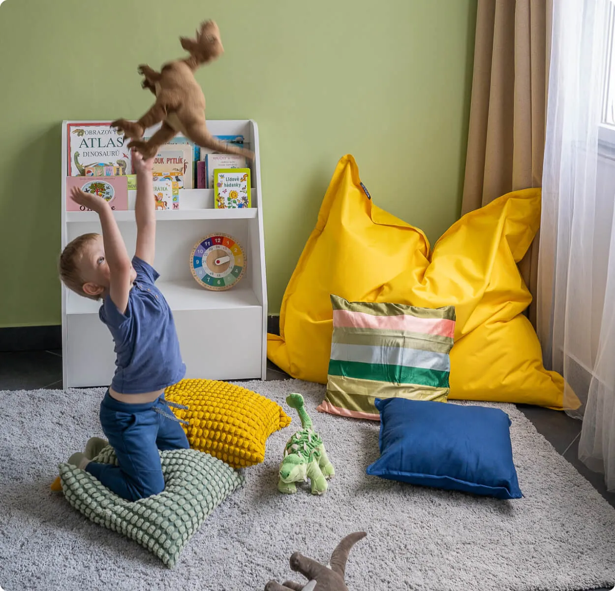 Chlapec klečí na polštářích a hází nahoru plyšového dinosaura v barevném pokoji pro děti.