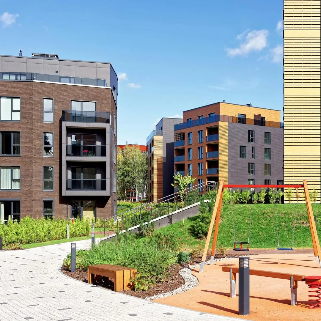 Modern residential buildings near a green playground with swings and a bench under a clear sky.
