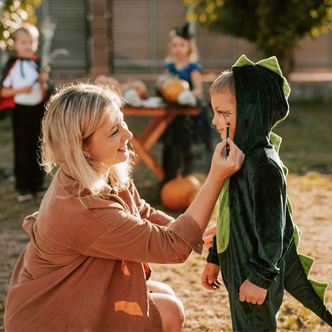 Žena maluje pavoučí pavučinu na tvář chlapce v kostýmu dinosaura na venkovním podzimním dni.
