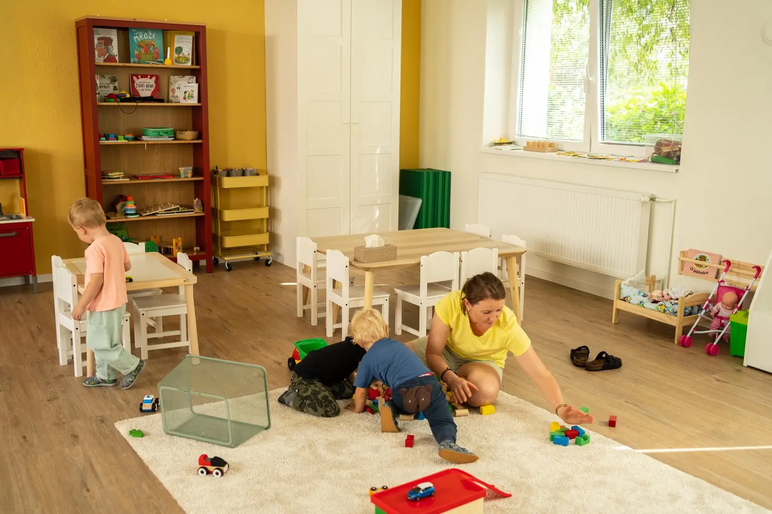 Děti a žena si hrají s kostkami a autíčky v jasné herně s dřevěným nábytkem.