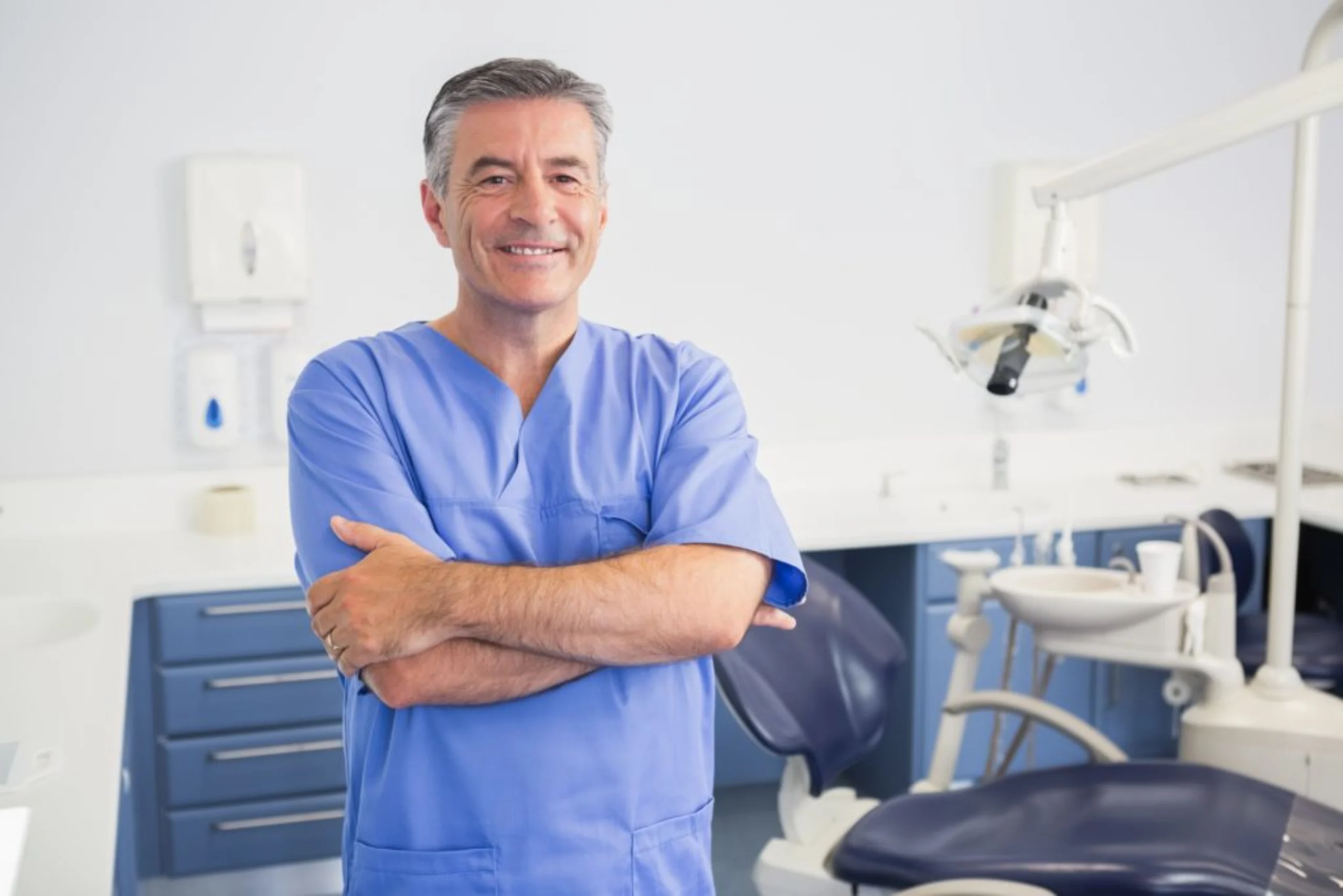 Smiling dentist in blue scrubs standing in dental office with equipment