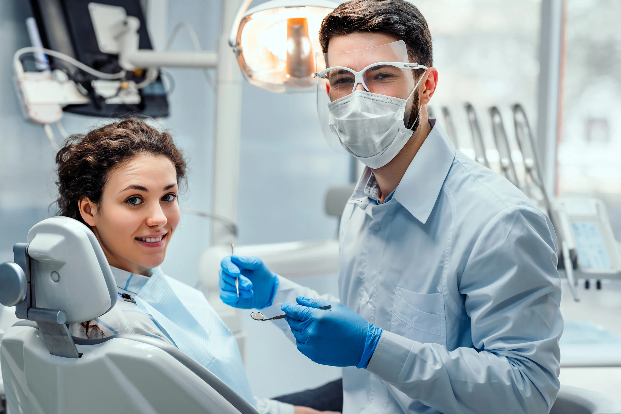 A young dentist working with a patient in the chair. The dentist is holding tools in his hands and both are smiling at the camera.