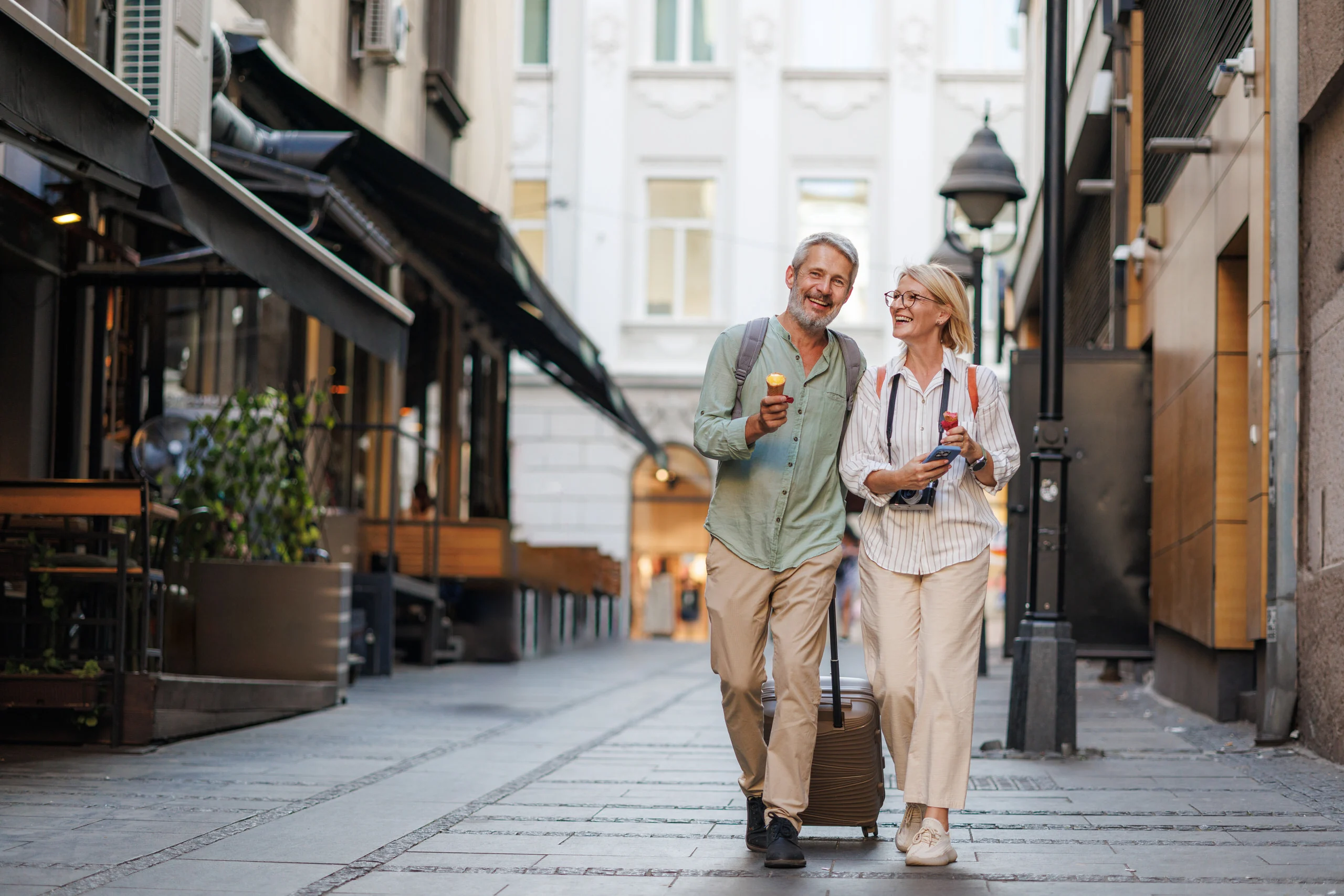 Smiling retired travelers walking together on city street with suitcase and ice cream