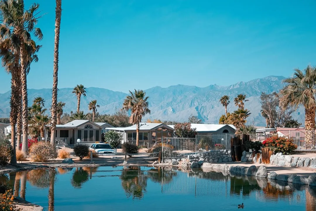 Desert mobile home community with pond, palm trees, and mountain backdrop under clear blue sky