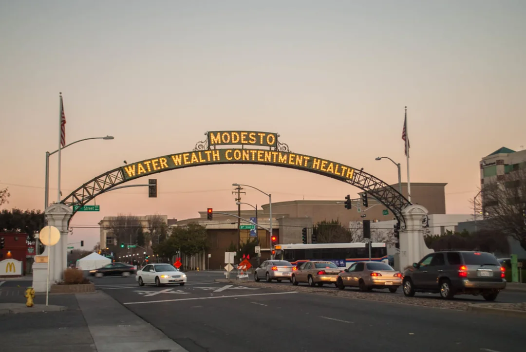 Modesto arch spanning street at dusk reading "WATER WEALTH CONTENTMENT HEALTH" with traffic and traffic lights below