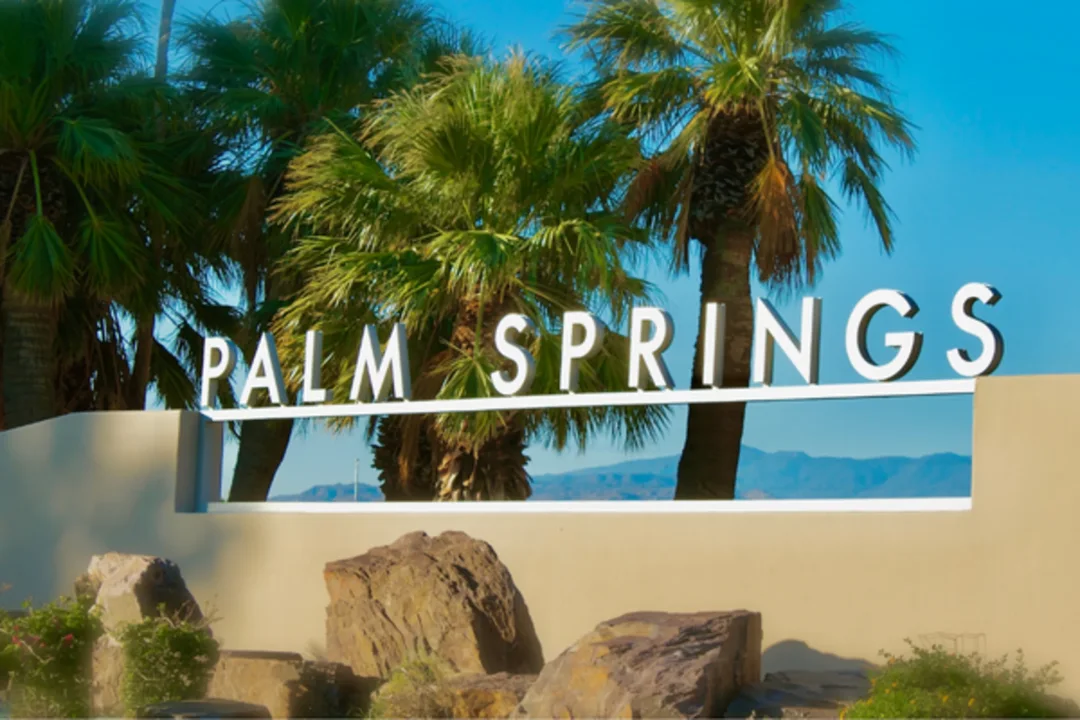 Palm Springs sign with palm trees and rocks against blue sky at city entrance