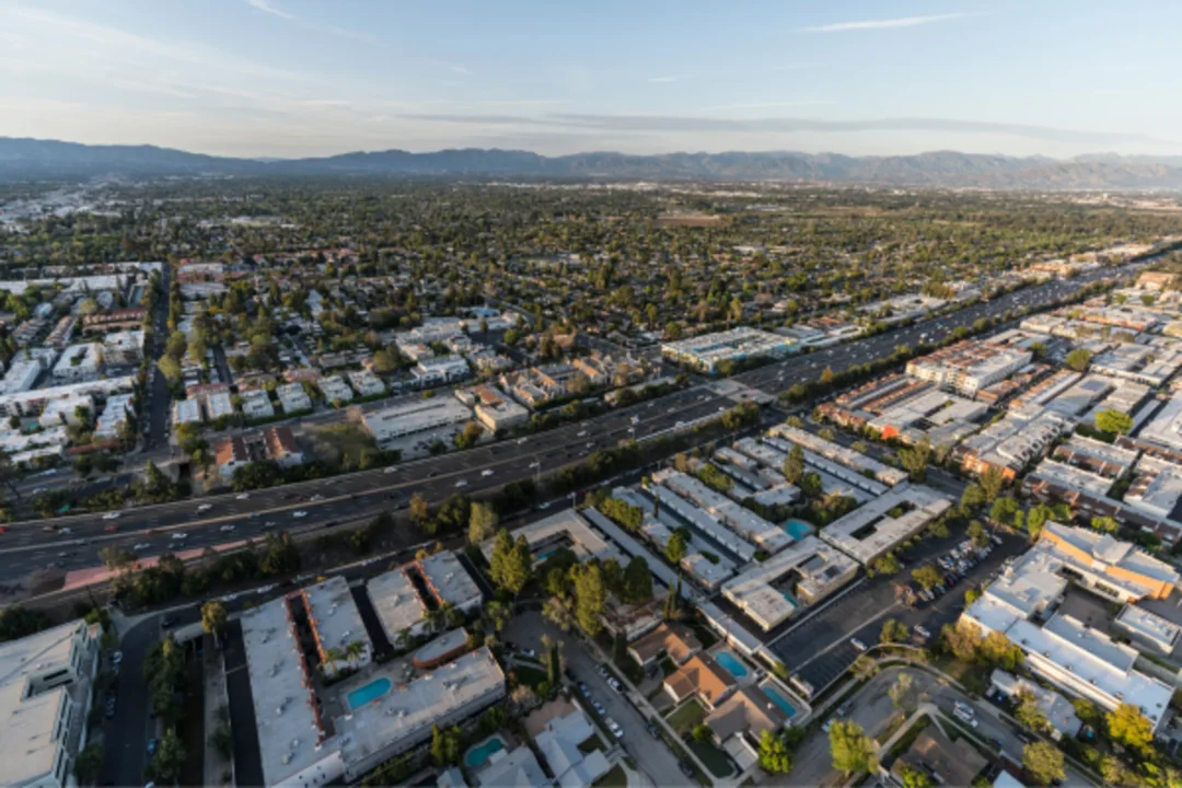 Aerial view of suburban sprawl with residential neighborhoods, commercial buildings, and a multi-lane highway leading to distant mountains.