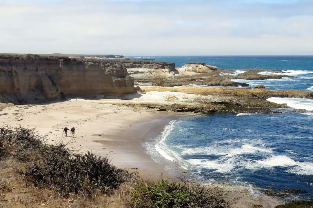 Rugged coastal cliff with sandy beach cove, rocky outcrops, and blue ocean waves with two people walking on shore.