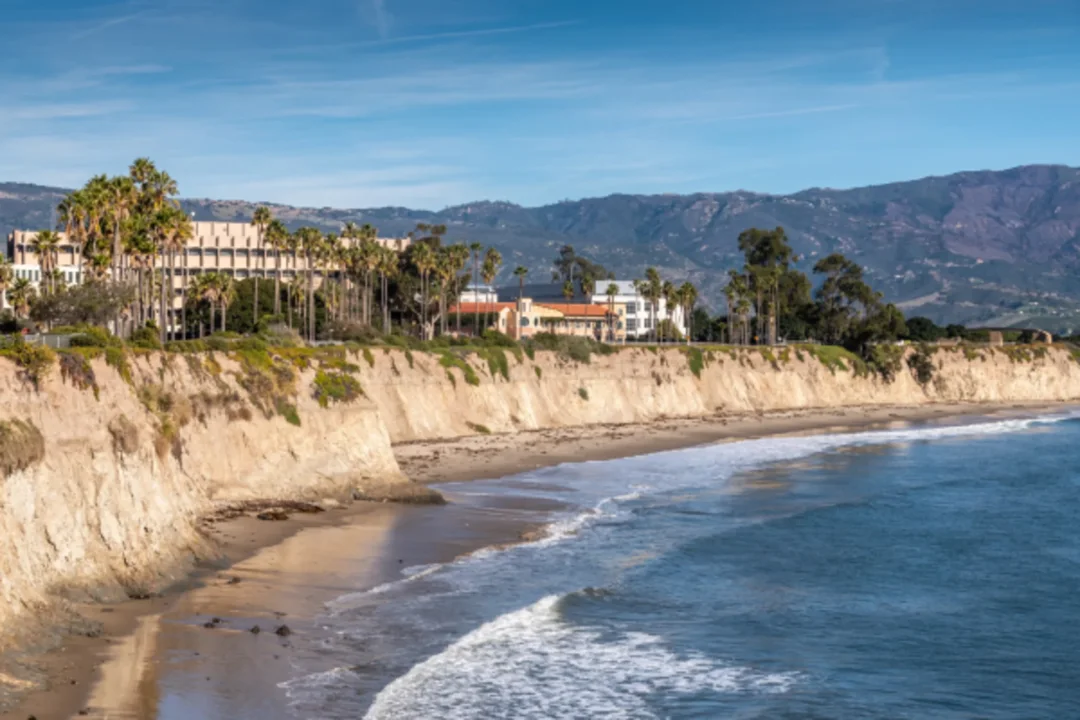 Coastal cliffs with palm trees and beachfront homes overlooking sandy beach and ocean waves, with mountains in background