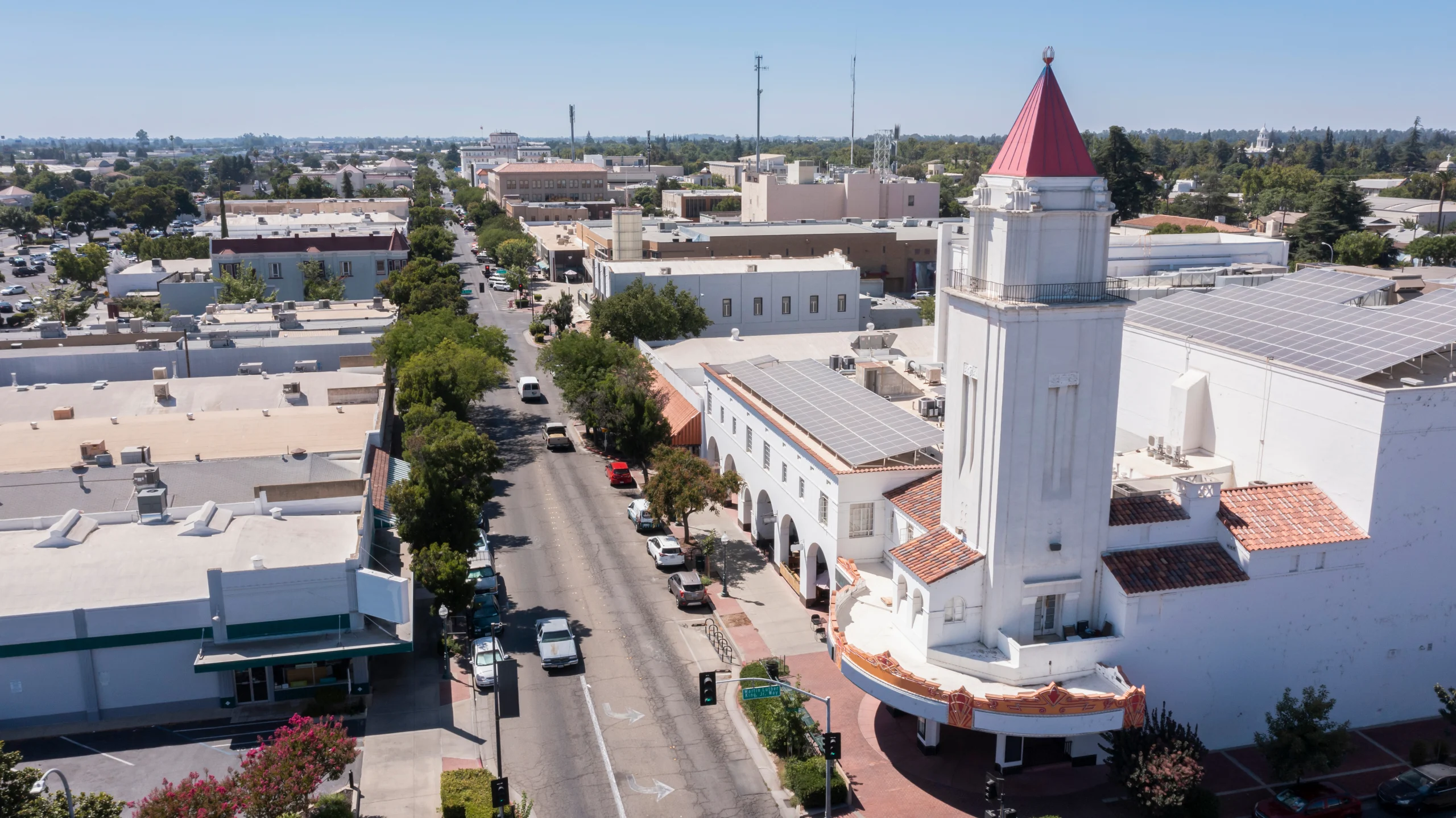 Aerial view of a small town main street with a white Spanish Colonial Revival church featuring a distinctive red-topped tower and solar panels on nearby buildings.