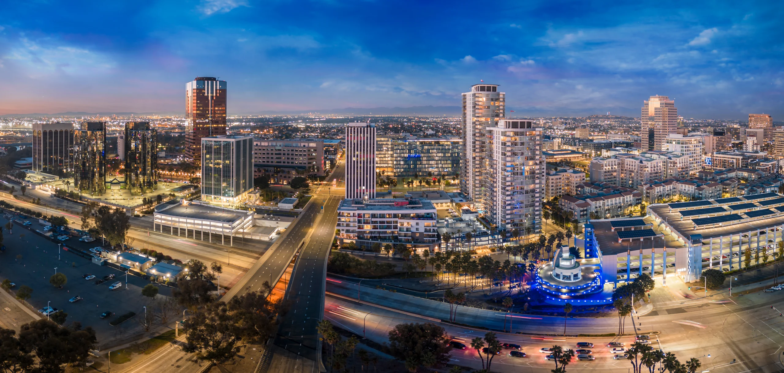 Aerial twilight view of Long Beach skyline with modern buildings, palm trees, and illuminated streets along the waterfront.