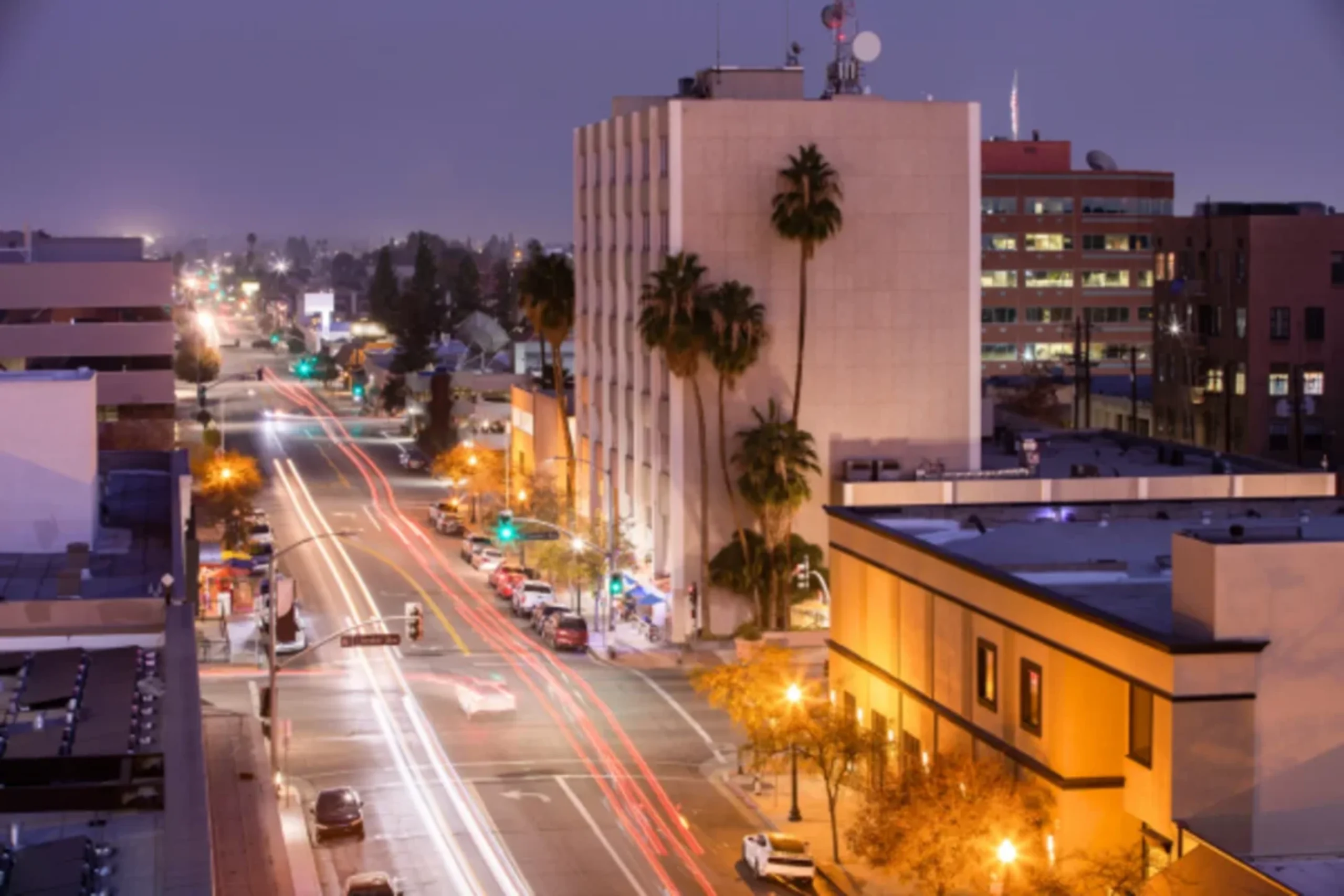 Urban street at dusk with light trails from traffic, palm trees, and illuminated buildings in a downtown area.