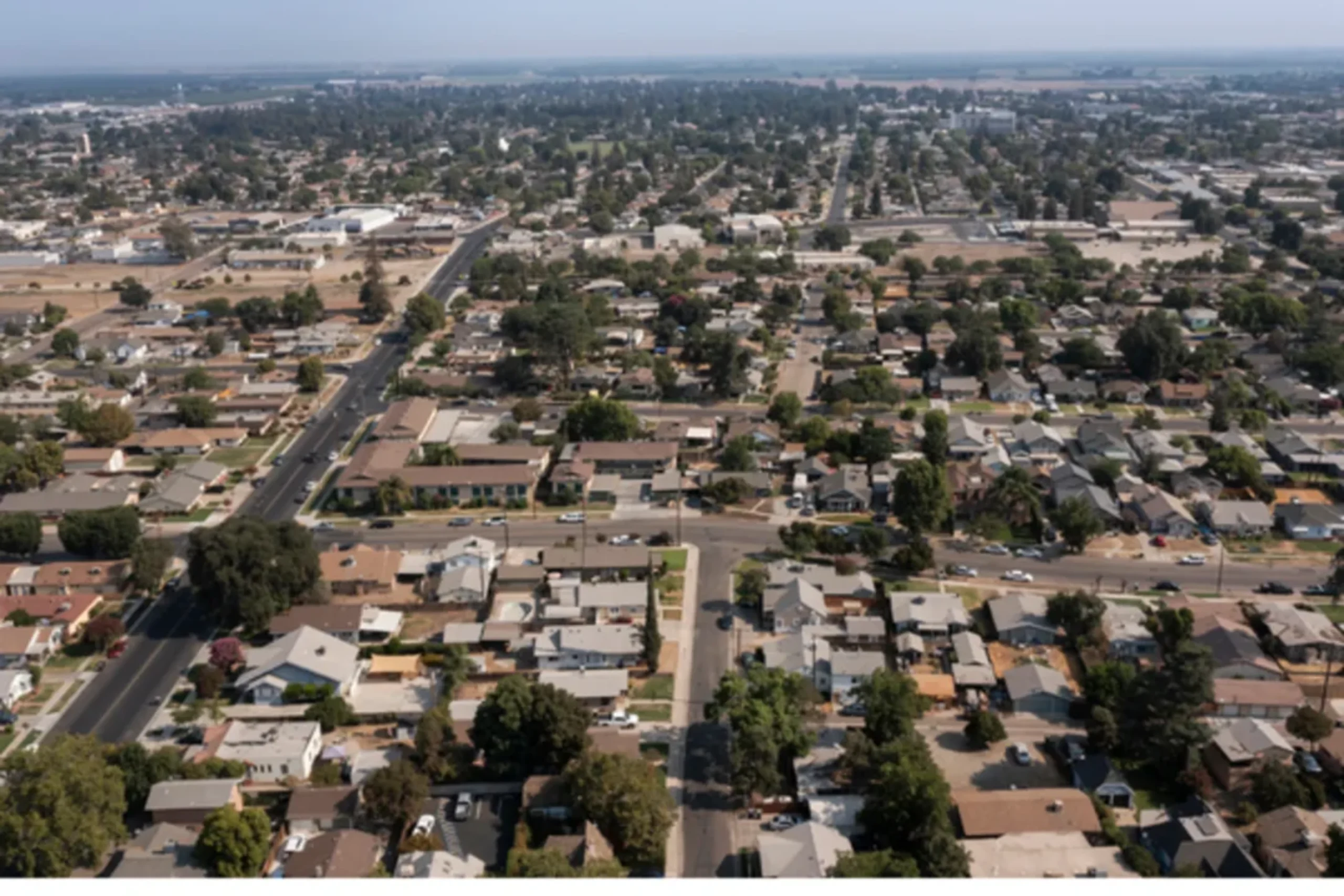 Aerial view of a suburban residential neighborhood with single-family homes, tree-lined streets, and distant farmland under hazy skies.