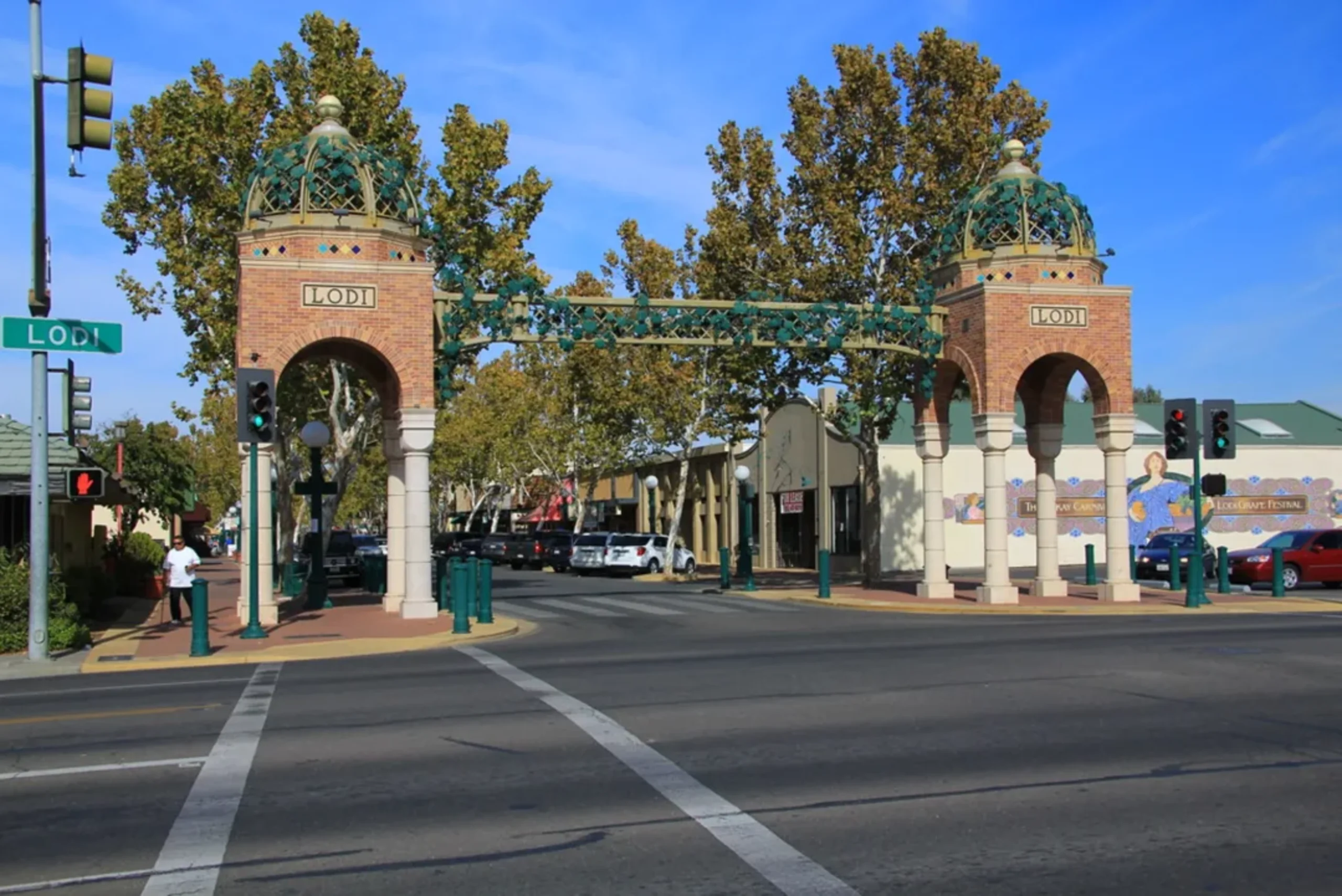 Ornate brick archway entrance to Lodi with decorative green domes and connecting bridge over a tree-lined street.