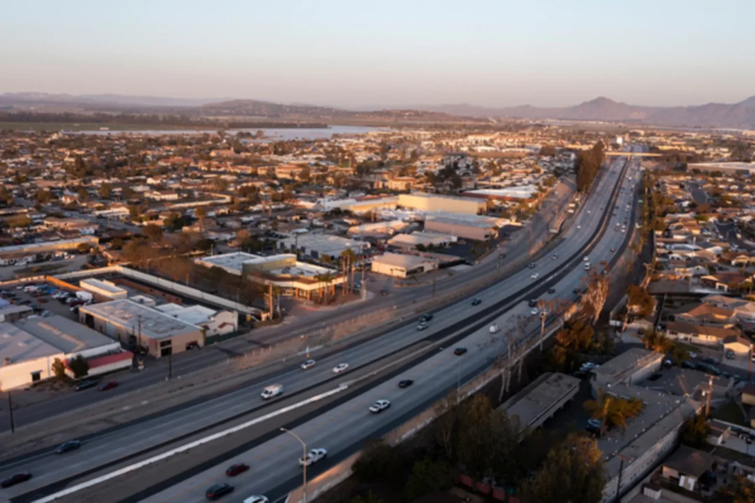 Aerial view of a multi-lane highway cutting through a suburban area with mountains and water in the distance at golden hour.