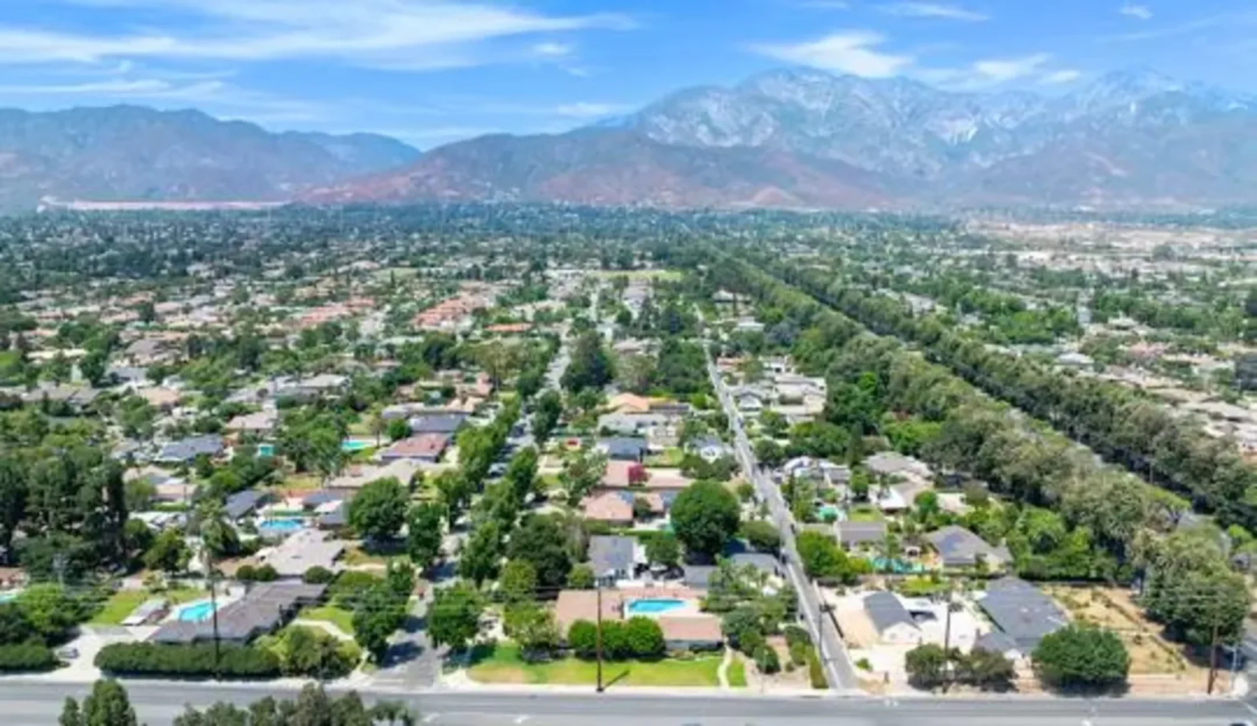 Aerial view of suburban neighborhood with tree-lined streets and snow-capped mountains in the background under blue sky.