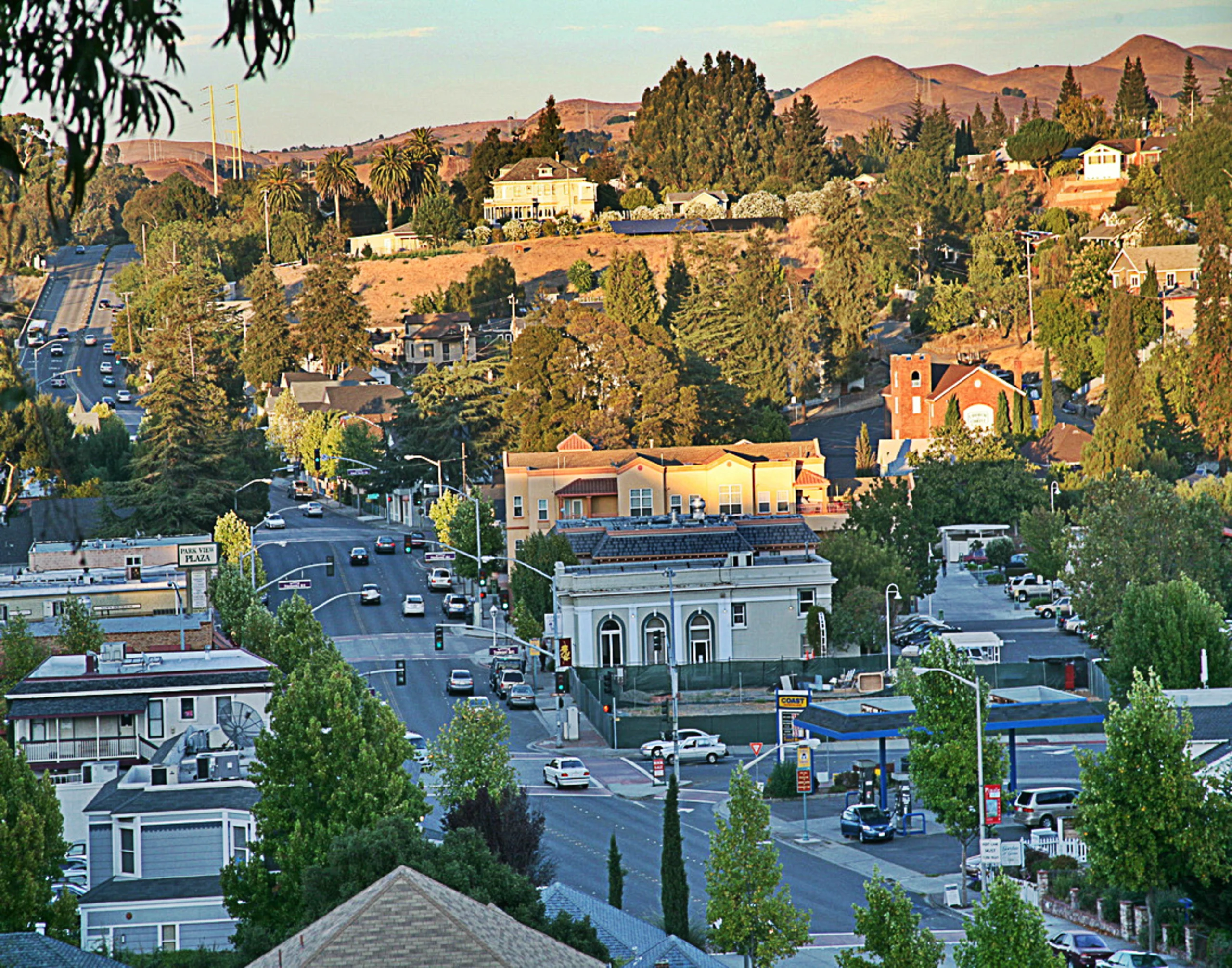Aerial view of a small hillside town with historic buildings, tree-lined streets, and brown hills in the background at golden hour.