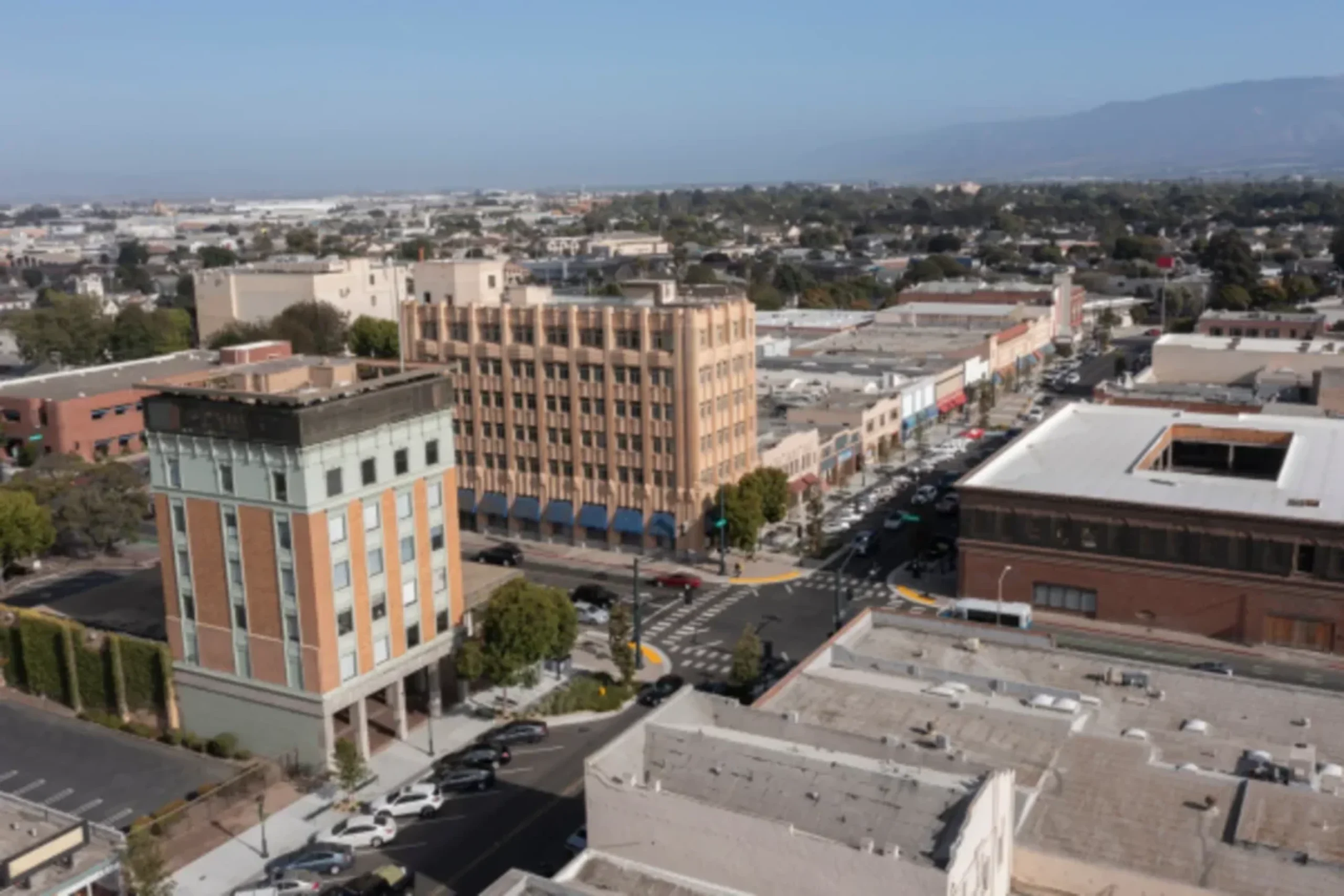 Aerial view of downtown urban area with multi-story buildings, streets with parked cars, and mountains in the background.