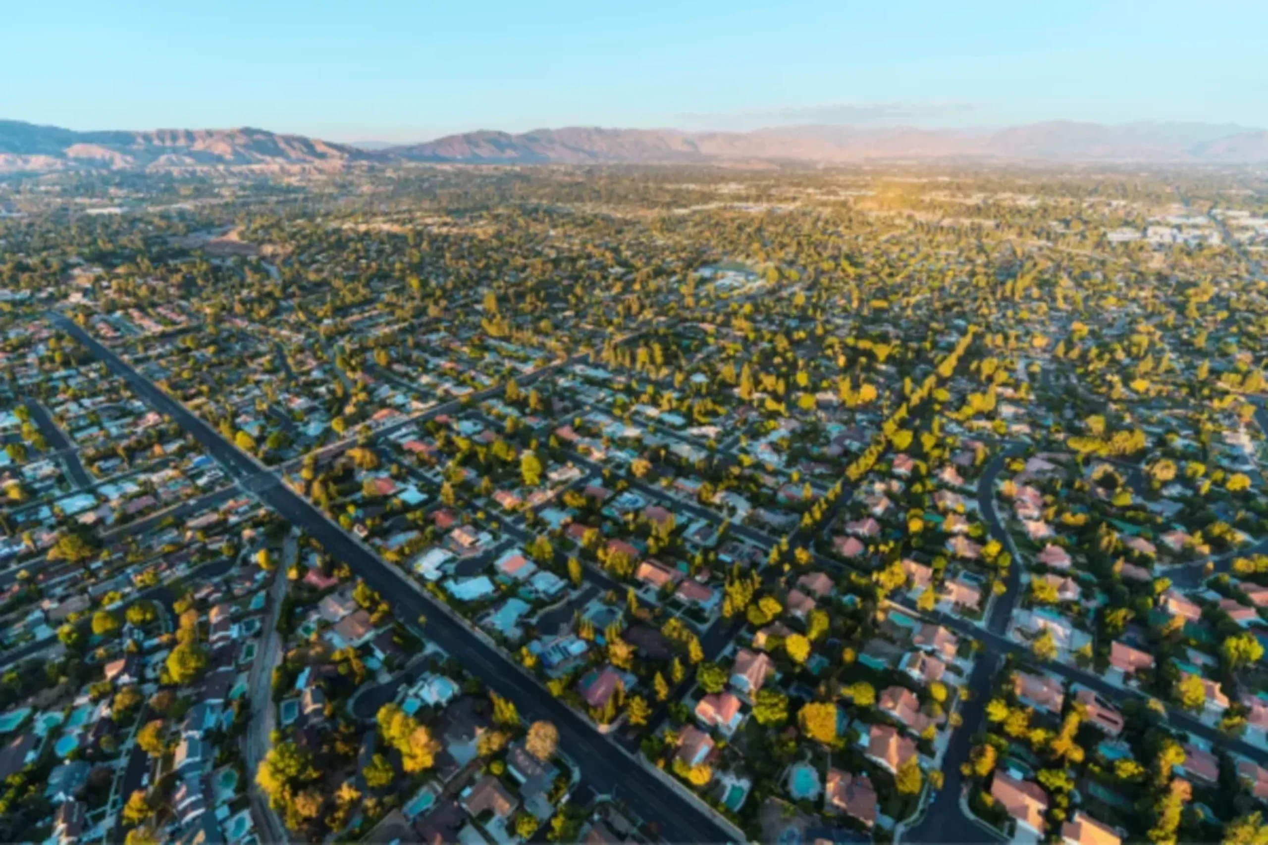 Aerial view of suburban neighborhood with tree-lined streets and houses stretching toward distant mountains under clear sky
