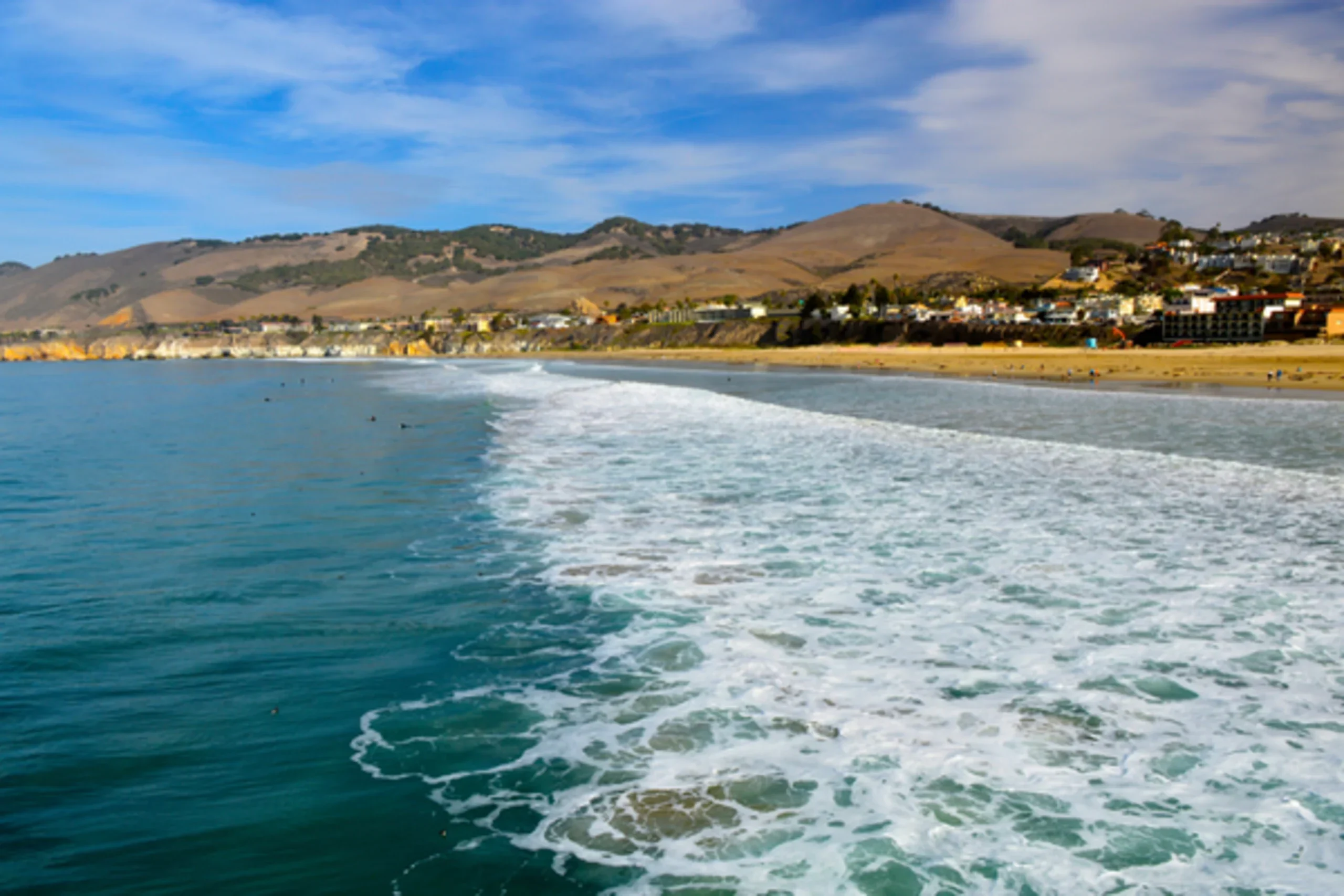 Ocean waves rolling toward a sandy beach with coastal town and golden hills under blue sky