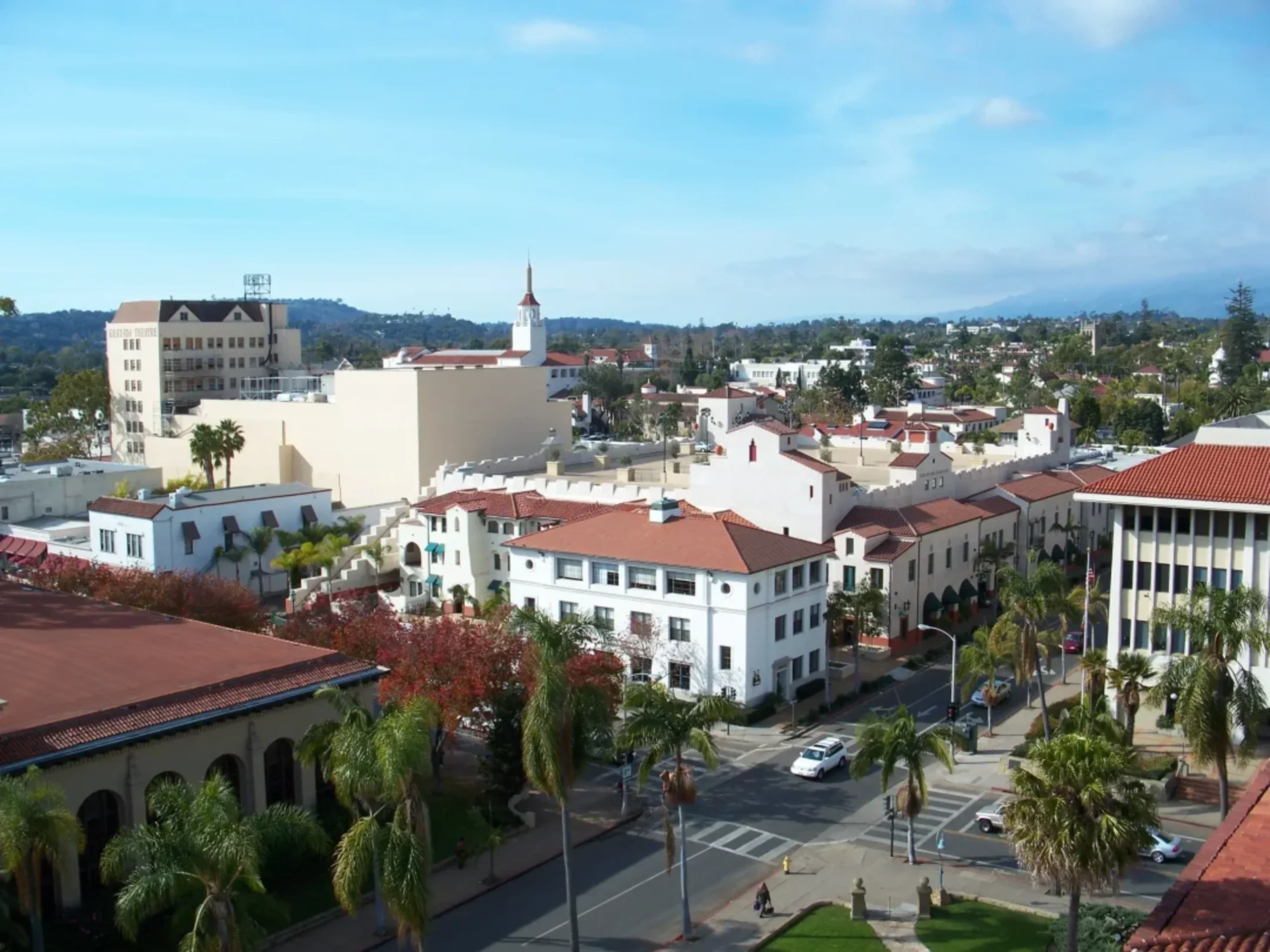 Aerial view of downtown Santa Barbara with white Spanish Colonial Revival buildings, red tile roofs, palm trees, and church steeple.