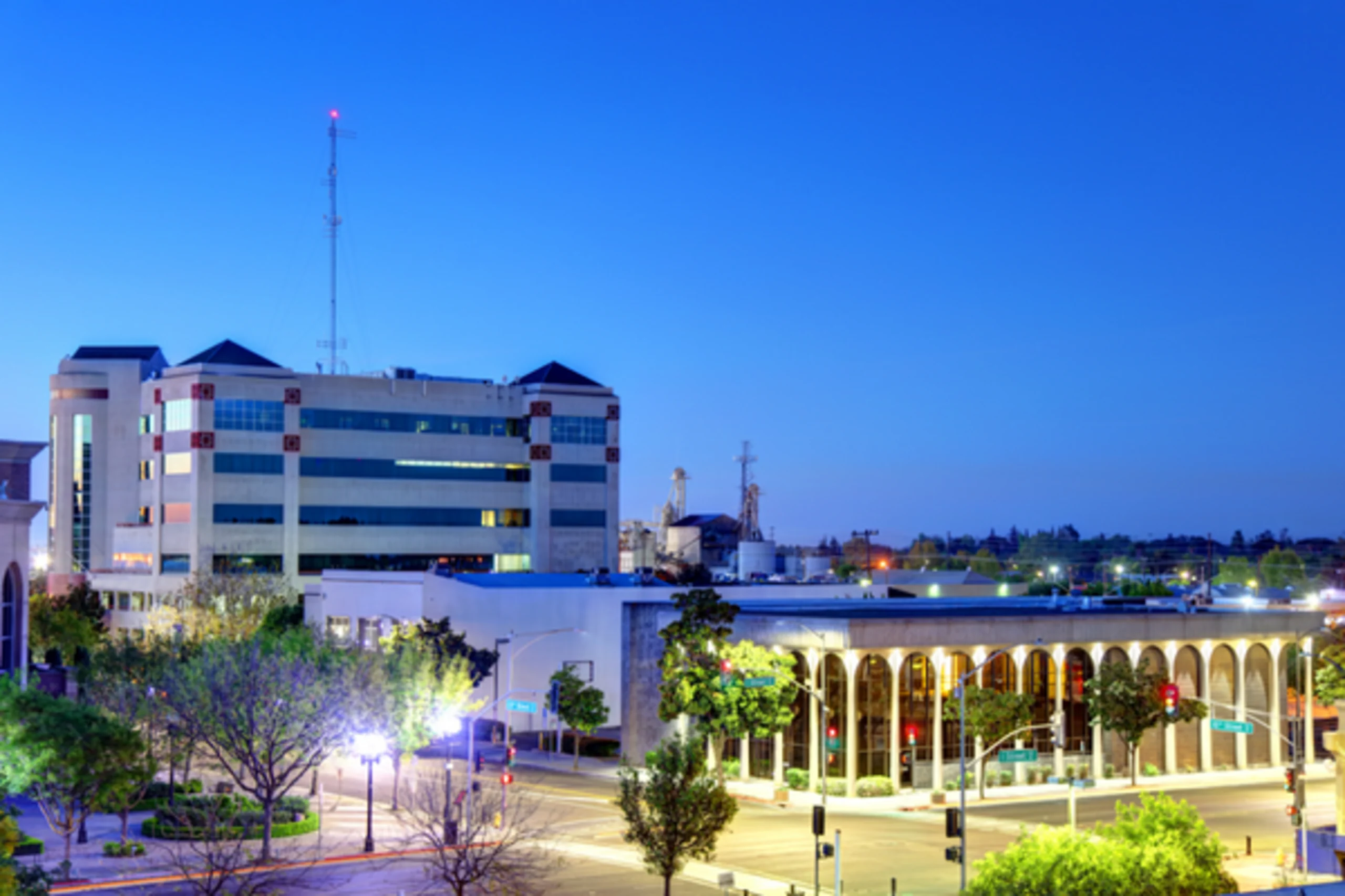 Urban skyline at dusk featuring modern buildings, a radio tower, and an illuminated colonnade structure with trees and streets below.
