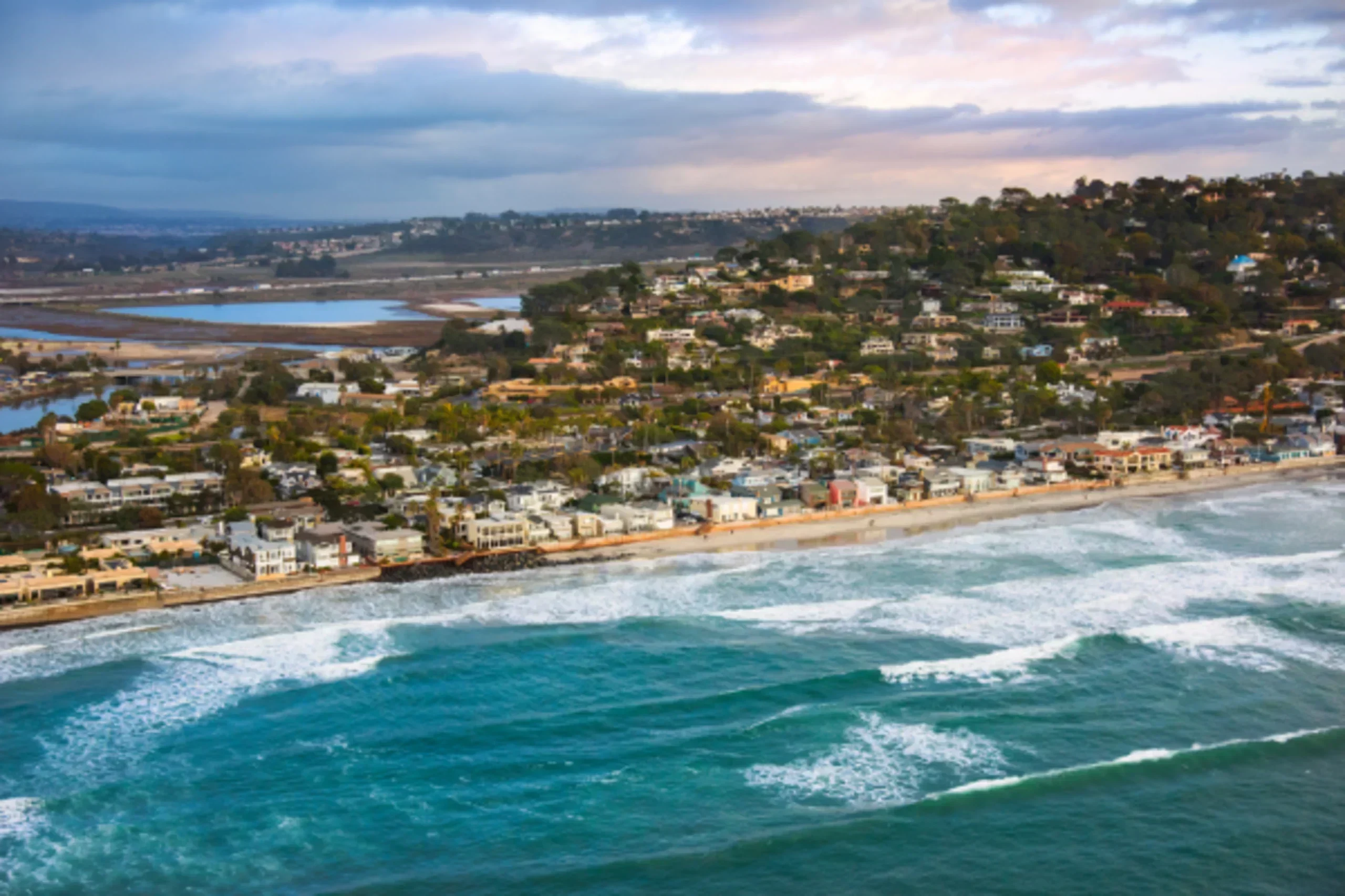 Aerial view of a coastal California town with beachfront homes, turquoise ocean waves, and a lagoon in the background at sunset.