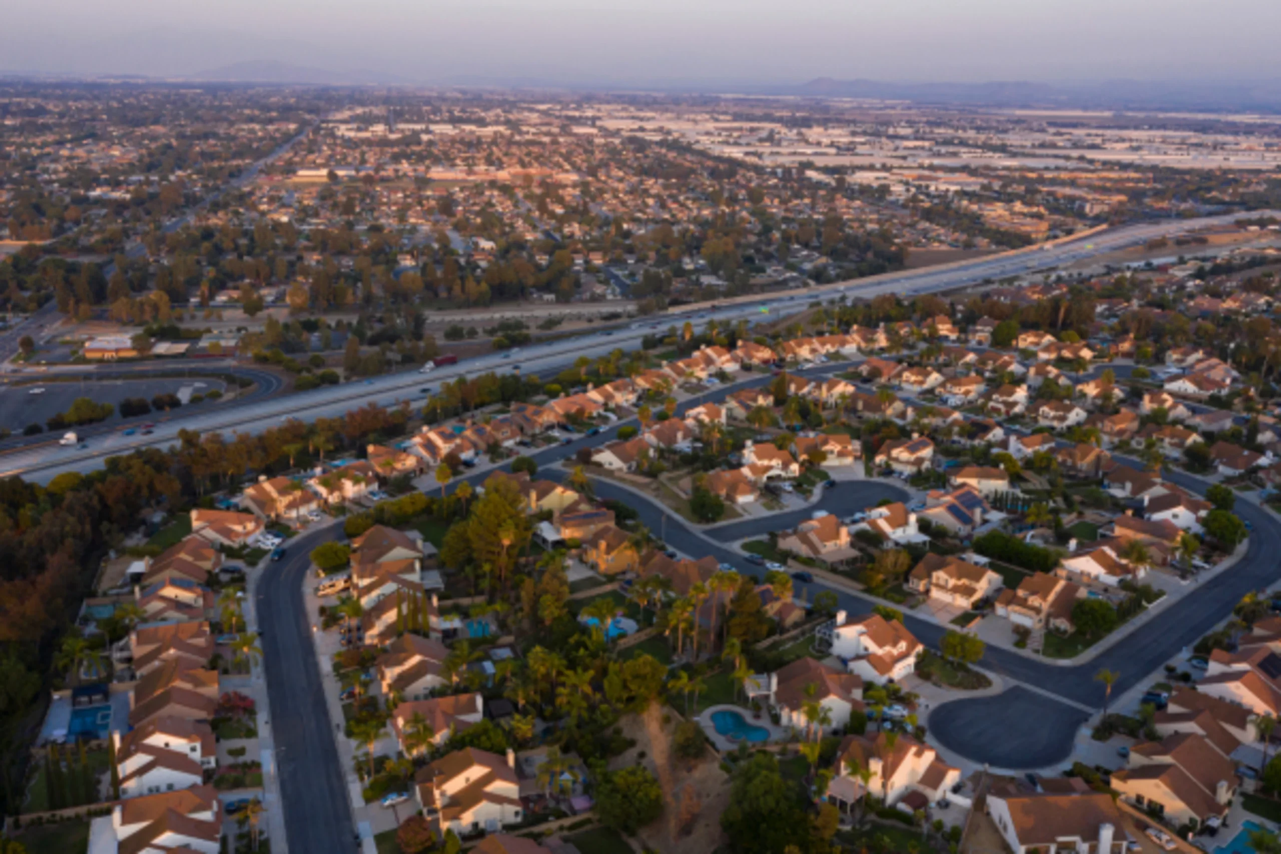 Aerial view of suburban residential neighborhood with curved streets, terracotta-roofed homes, and a highway in the background at dusk.