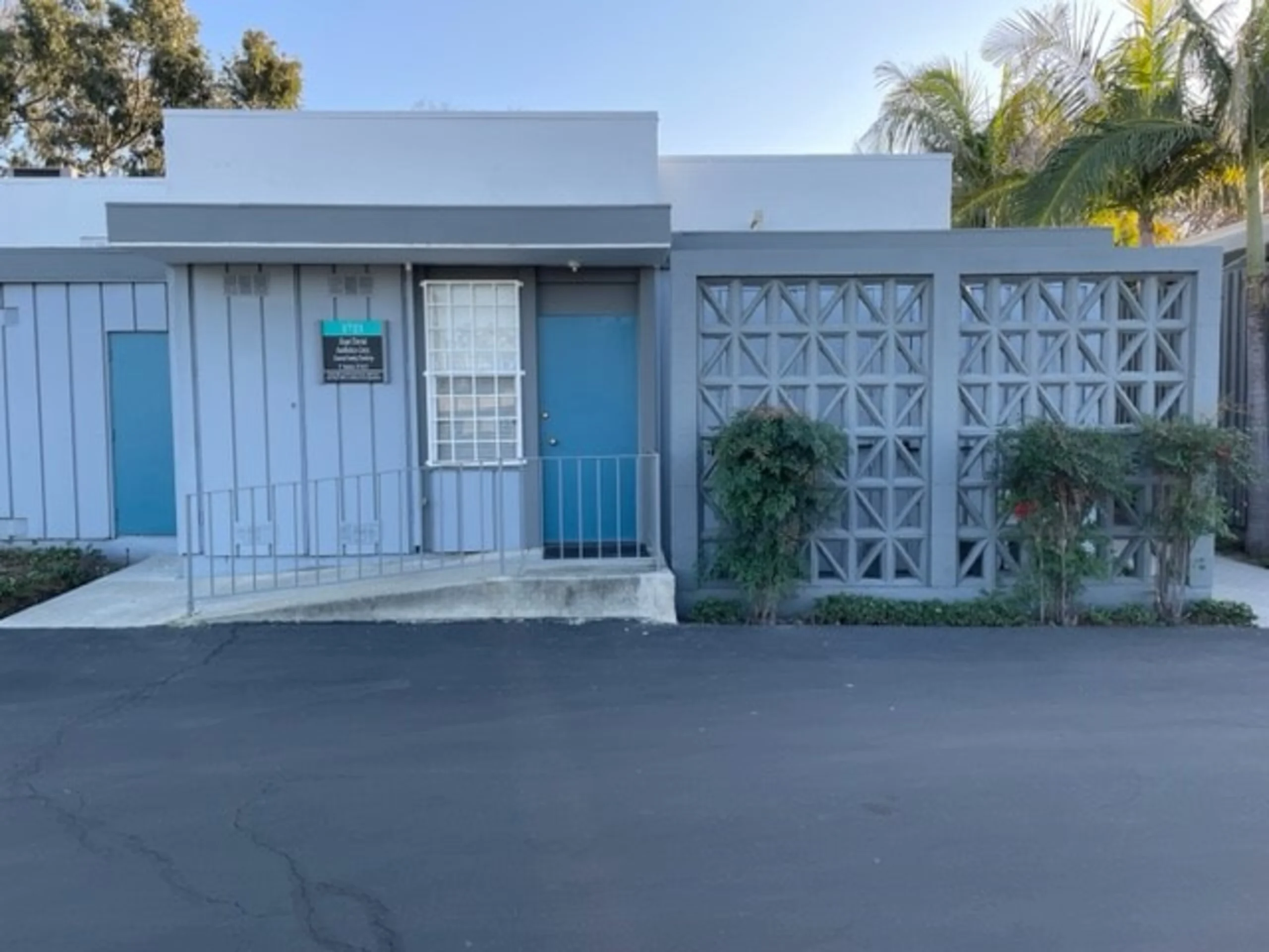 Modern white and gray building with decorative concrete block walls, palm trees, and blue doors in parking lot setting.