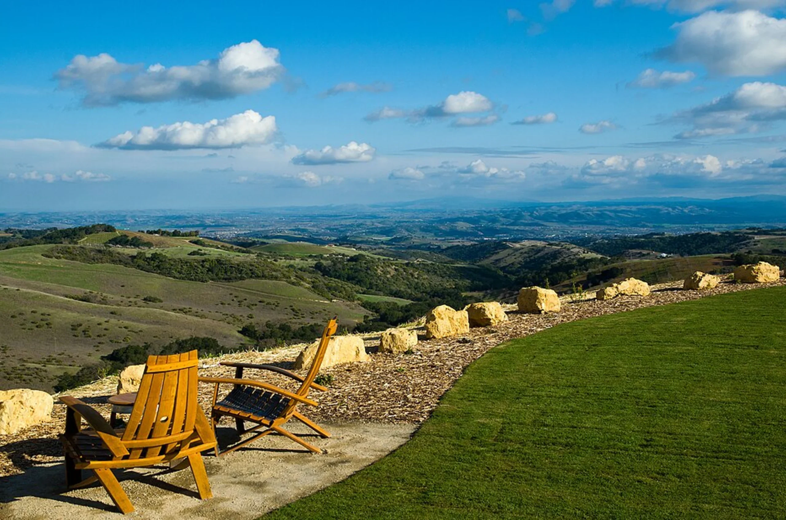 Two wooden chairs on a hilltop overlooking rolling green valleys under a blue sky with white clouds.