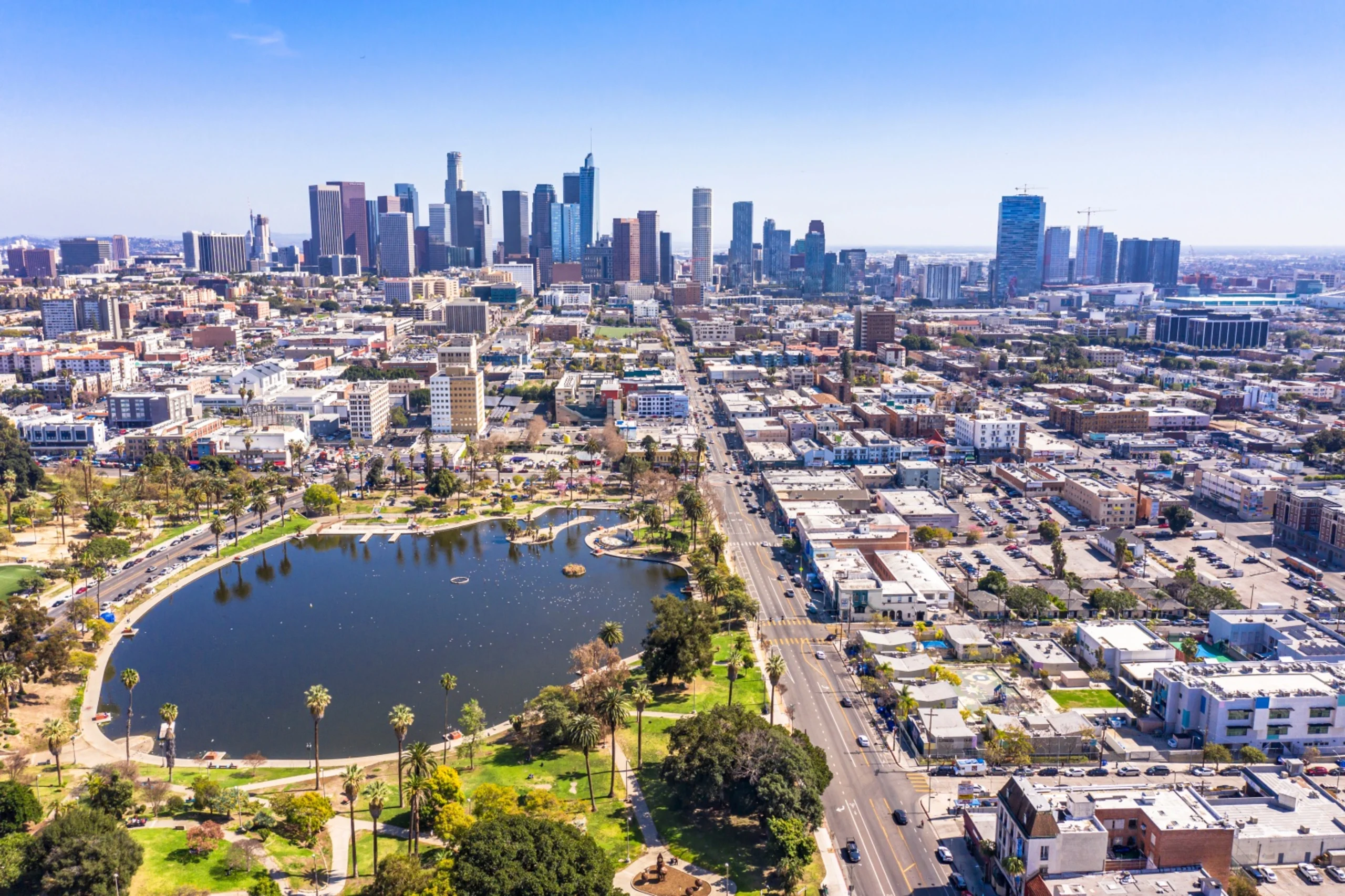 Aerial view of downtown Los Angeles skyline with MacArthur Park lake in foreground, surrounded by palm trees and urban neighborhoods.