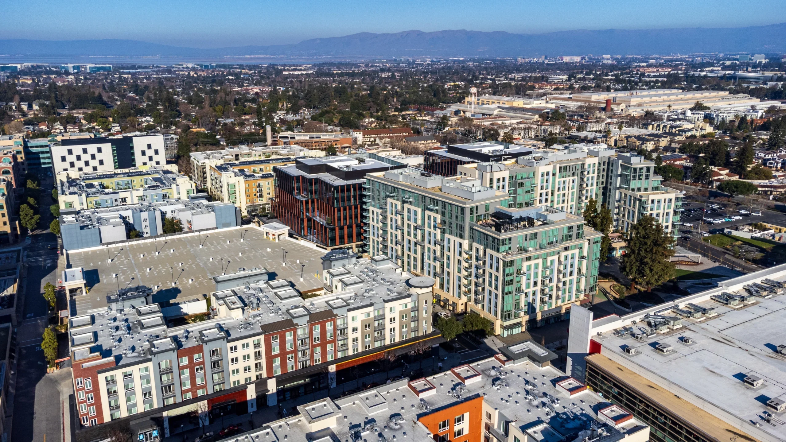 Aerial view of modern mixed-use development with colorful mid-rise buildings, surrounded by urban sprawl and mountains in the distance.