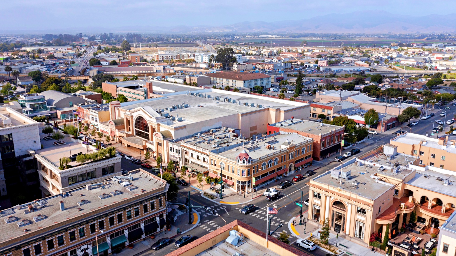 Aerial view of downtown urban area with multi-story buildings, streets with parked cars, and mountains in the background.