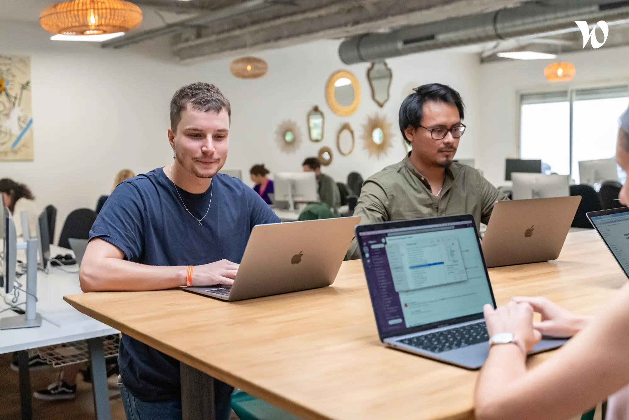 Trois personnes travaillent sur leurs ordinateurs portables autour d'une grande table en bois dans un bureau moderne.