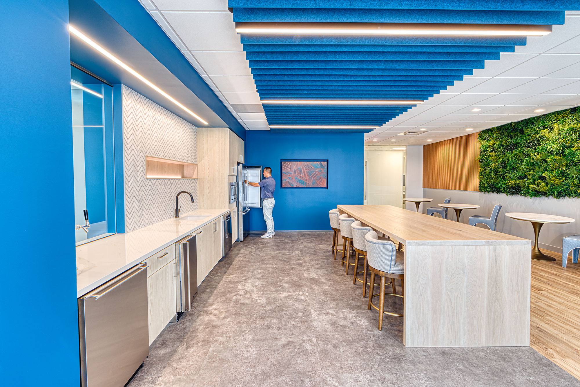 Breakroom and lounge by Ossa Studio with long wood table, person at fridge, blue baffles and a living green wall; biophilic workplace interior.