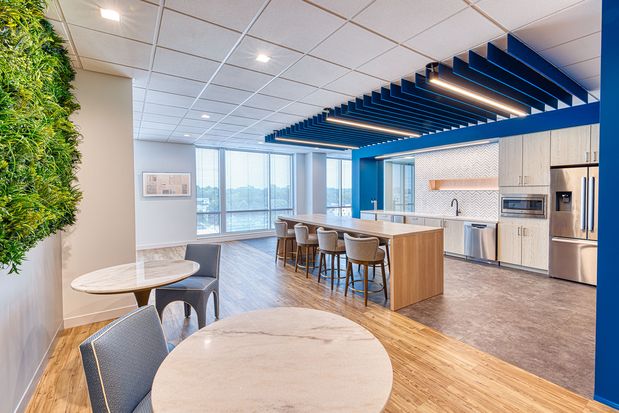 Café-style break area by Ossa Studio with green wall, bar-height island, seating and large windows; biophilic, light-wood office interior design.