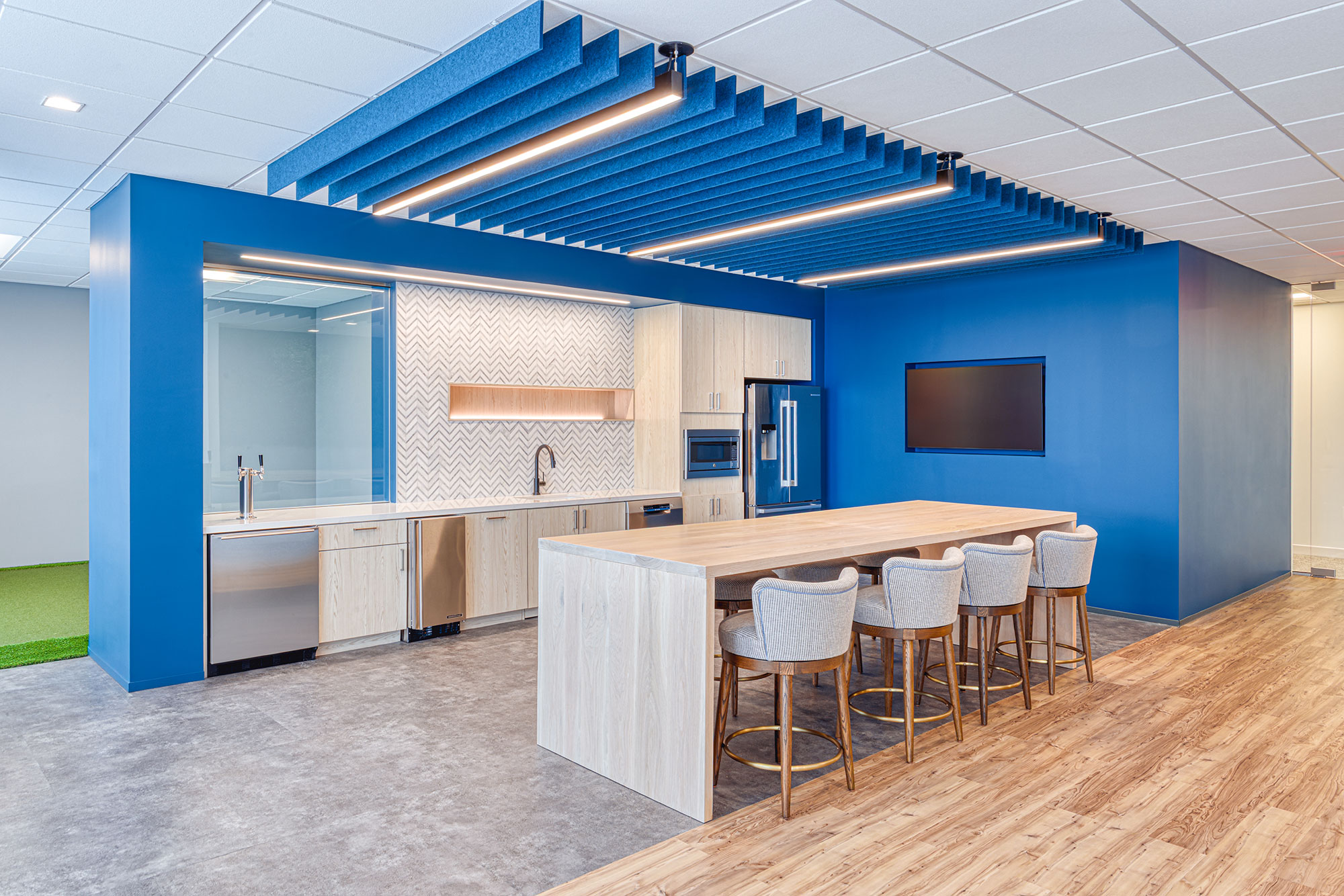 Office kitchen by Ossa Studio with light wood island, stainless appliances, chevron backsplash, and blue acoustic baffles; modern workplace design.