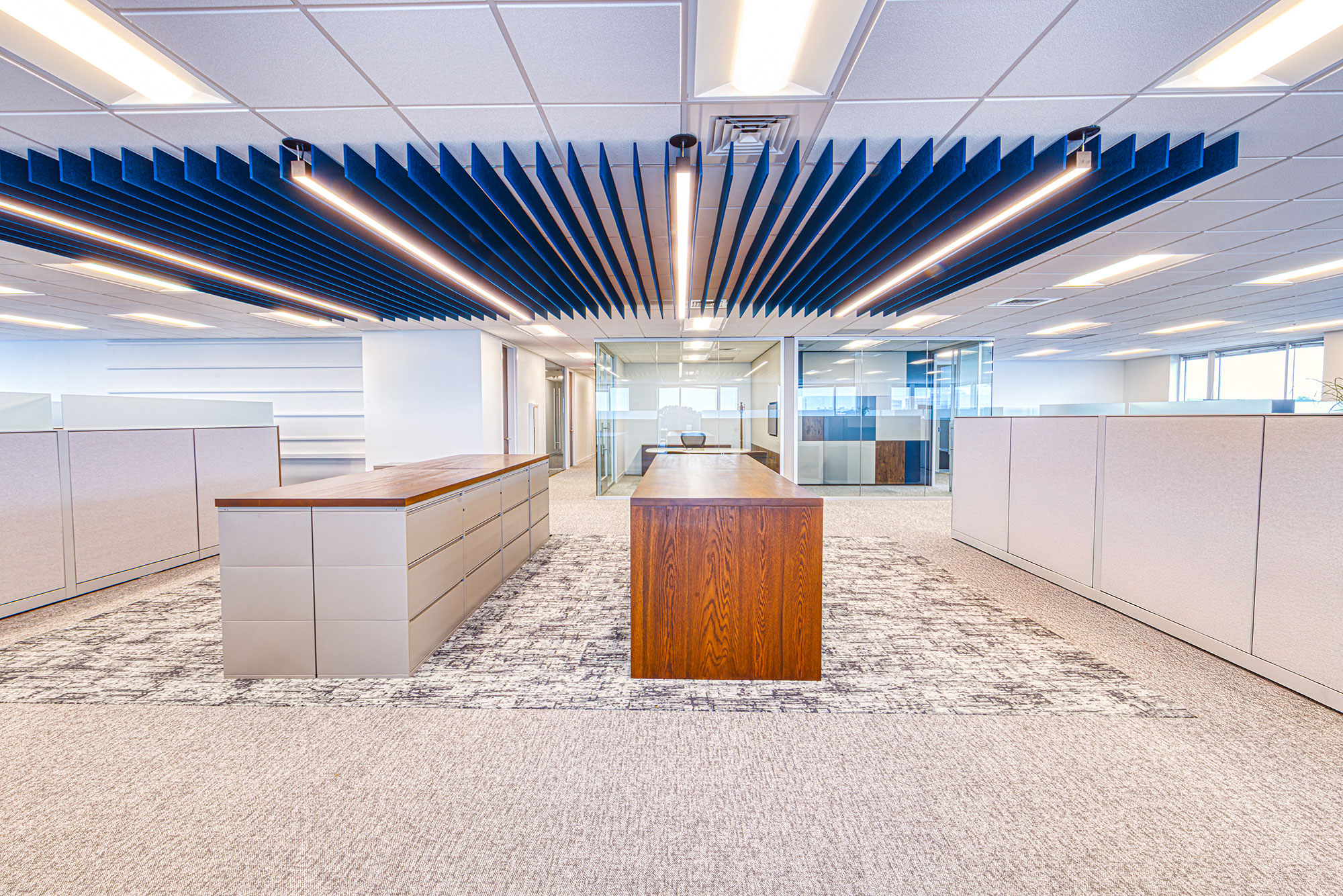 Open office corridor by Ossa Studio with storage islands, wood work table, glass rooms and continuous blue ceiling baffles; workplace interior design.
