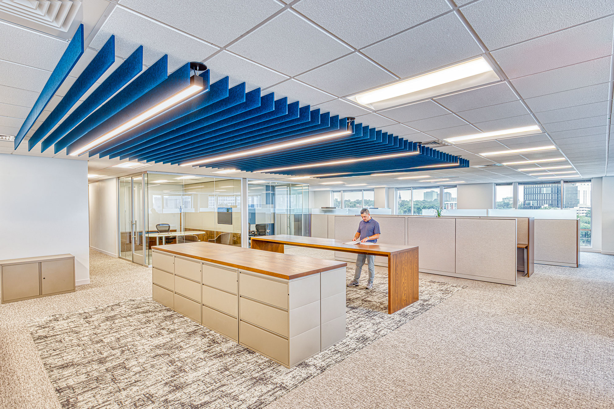 Open office by Ossa Studio with glass meeting rooms, workstations, linear lighting and blue acoustic baffles; collaborative workspace design.