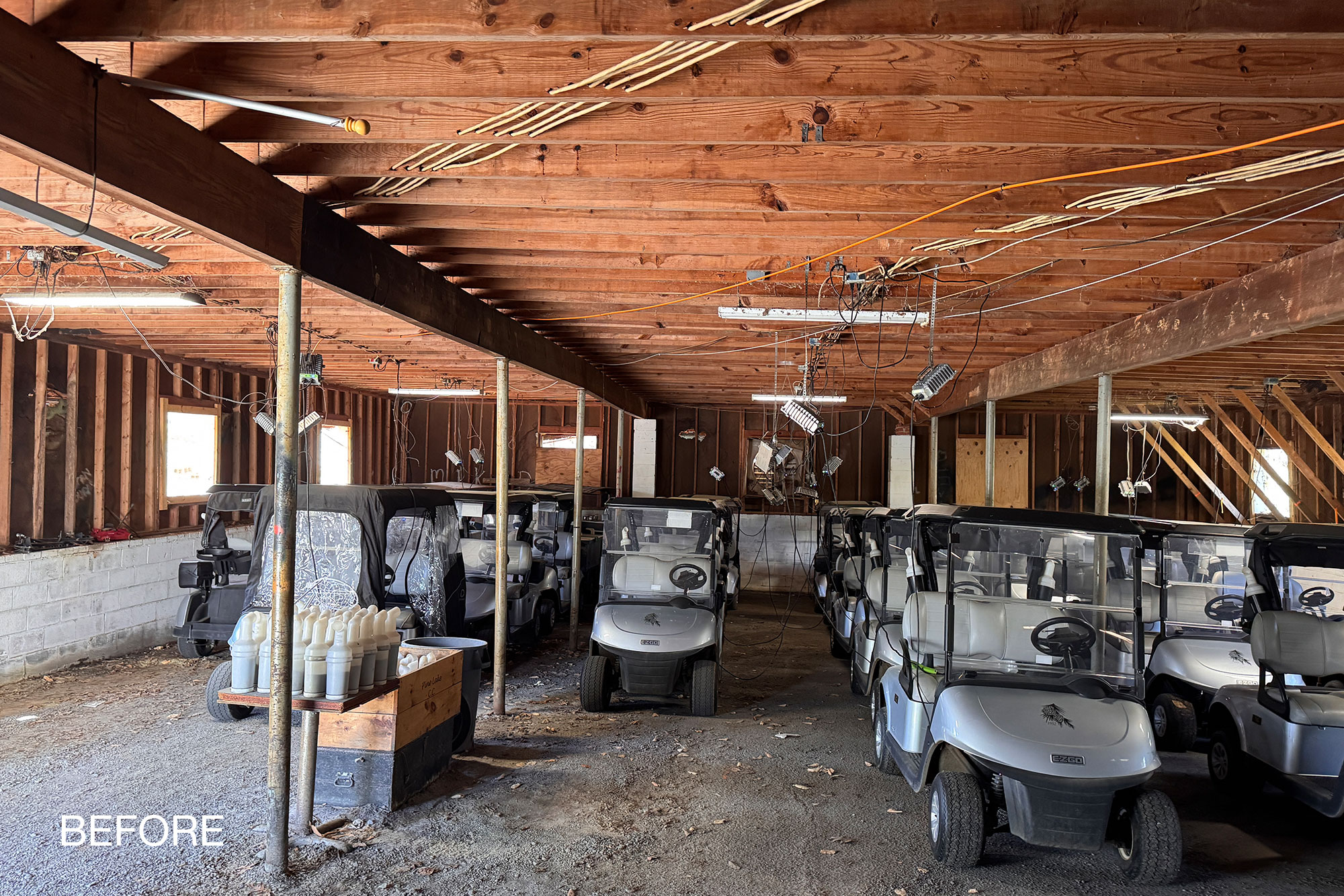 Pine Lake cart barn interior before renovation, with exposed wiring, steel pipe columns and golf carts on gravel.