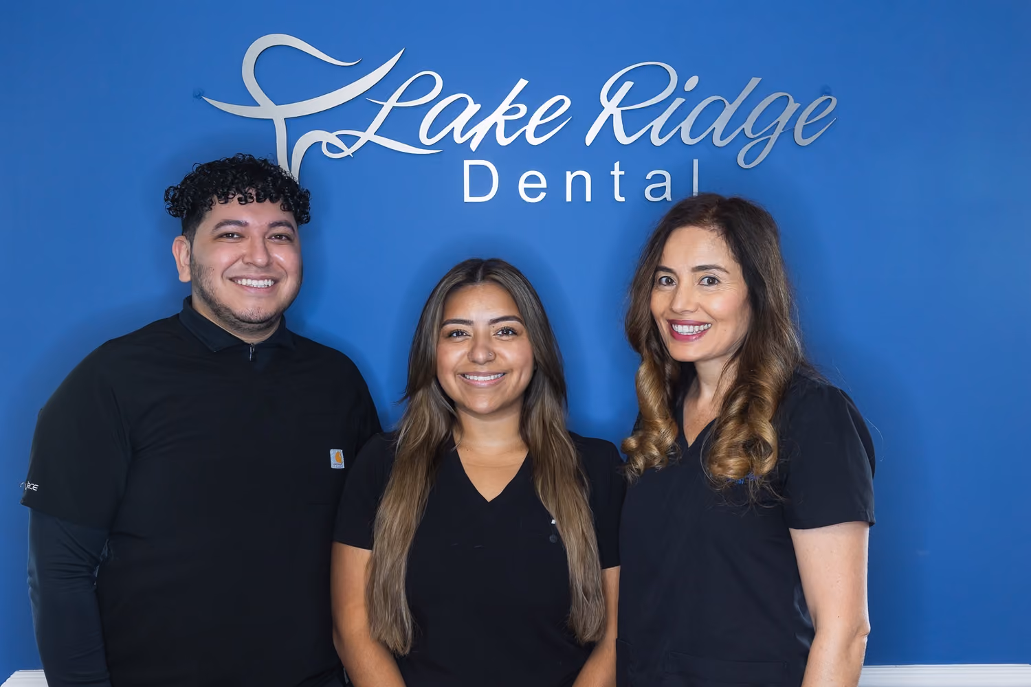 Three smiling dental professionals standing in front of a blue wall with 'Lake Ridge Dental' sign.