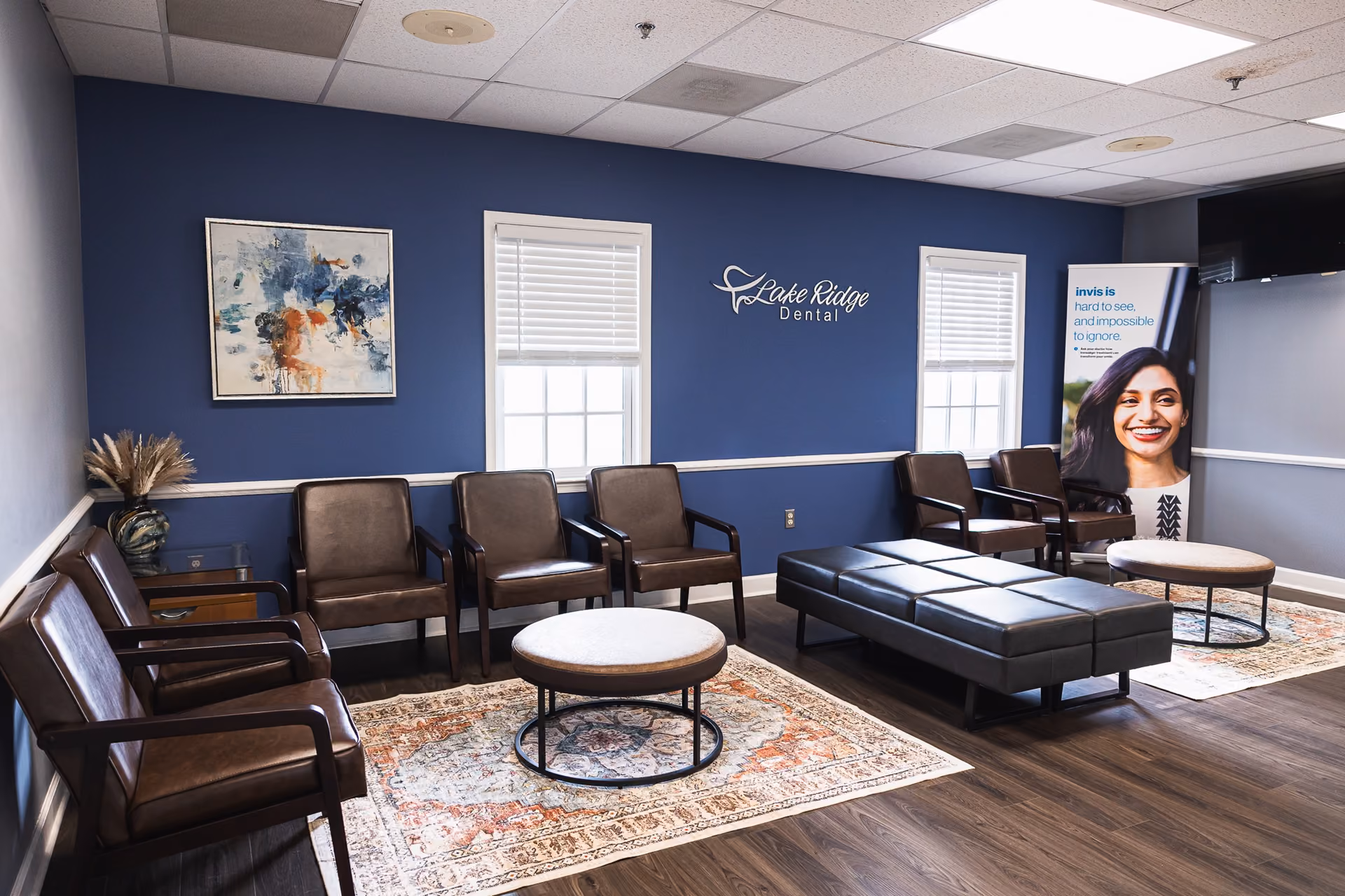 Modern dental office waiting room with brown leather chairs, blue walls, two windows, round tables, area rugs, and a sign reading 'Lake Ridge Dental'.
